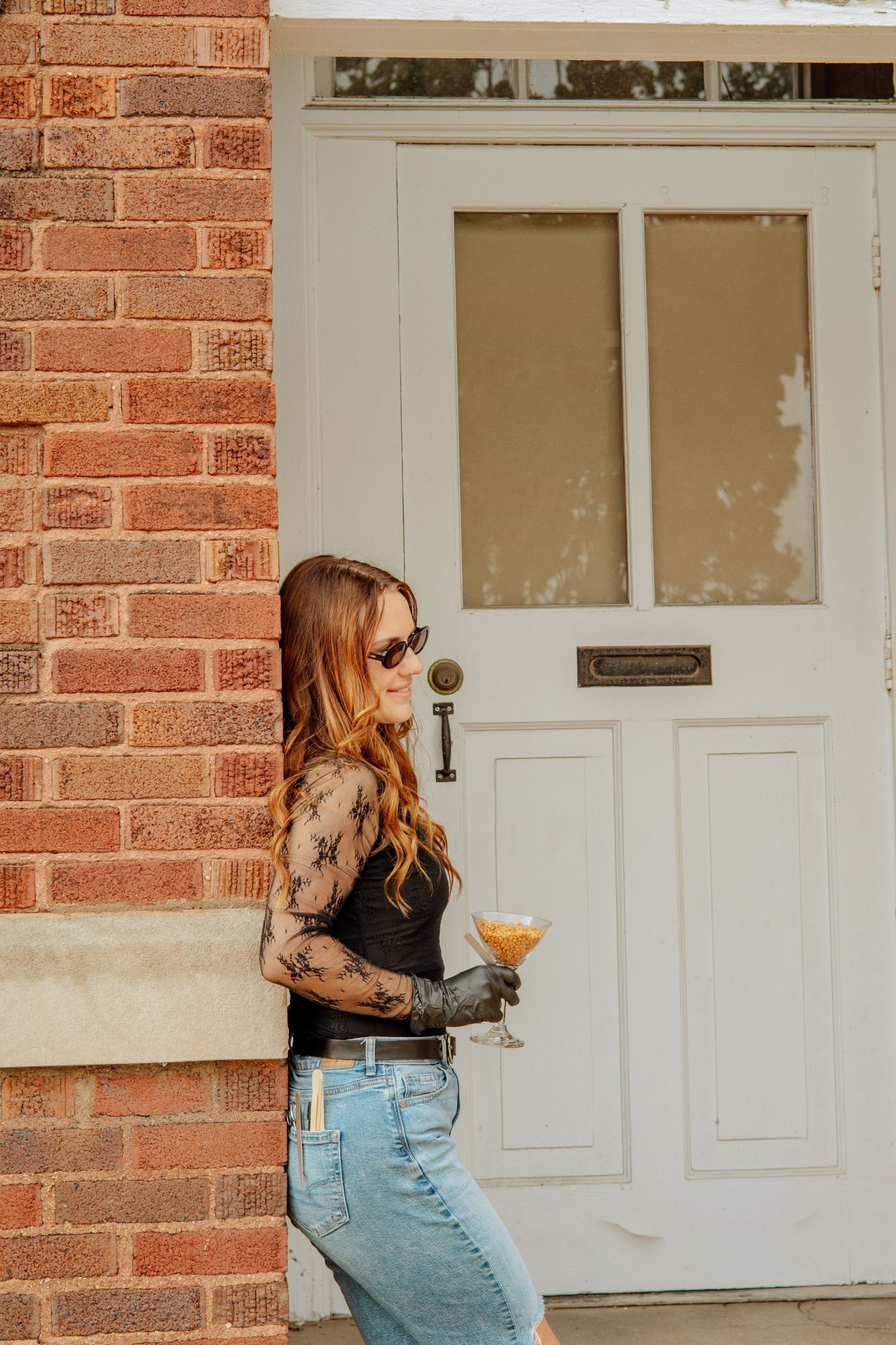 Stylish redhead woman in black lace top holding cocktail glass leaning against a brick wall outdoors.