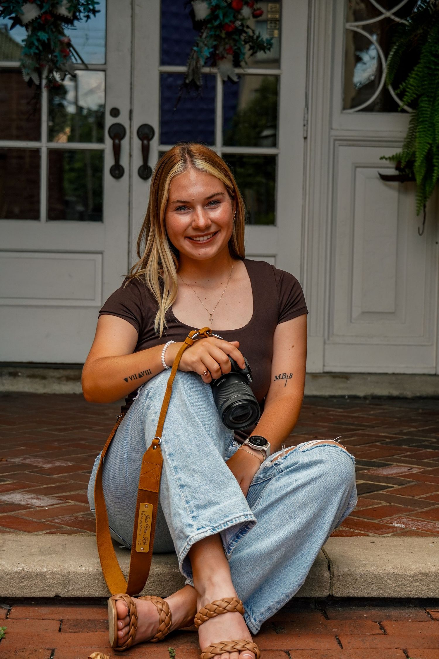 Young woman sitting on brick steps holding a camera with a tan strap, wearing jeans and a brown top.