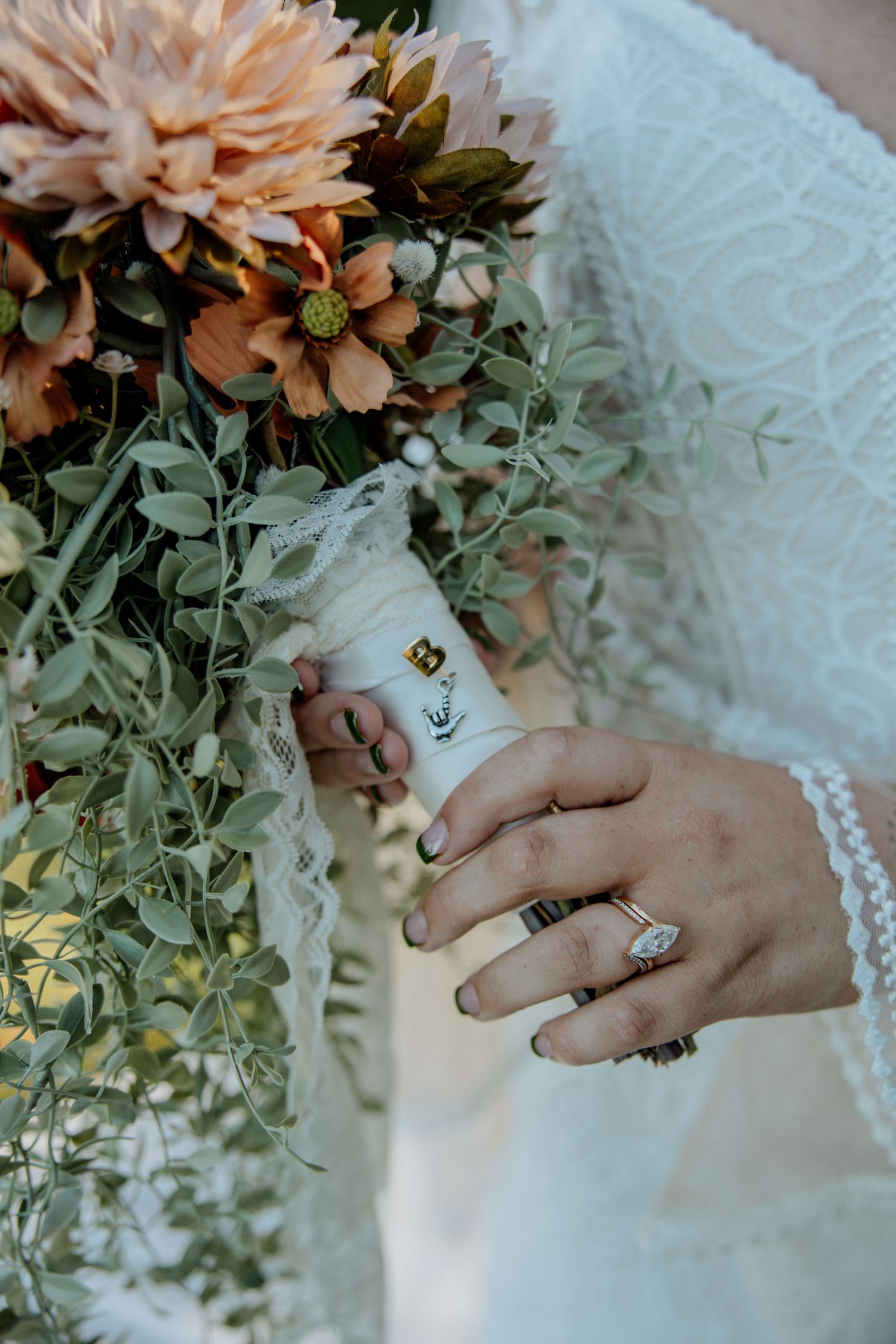 Bride in lace dress holding a bouquet of peach dahlias and greenery, showcasing a diamond ring on her finger.