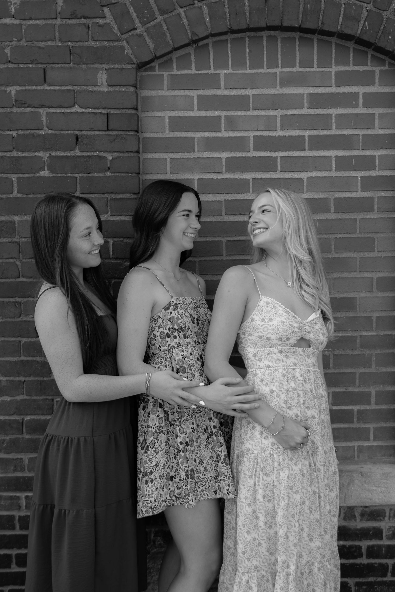 Three smiling women in summer dresses standing together against a brick wall in black and white photo.