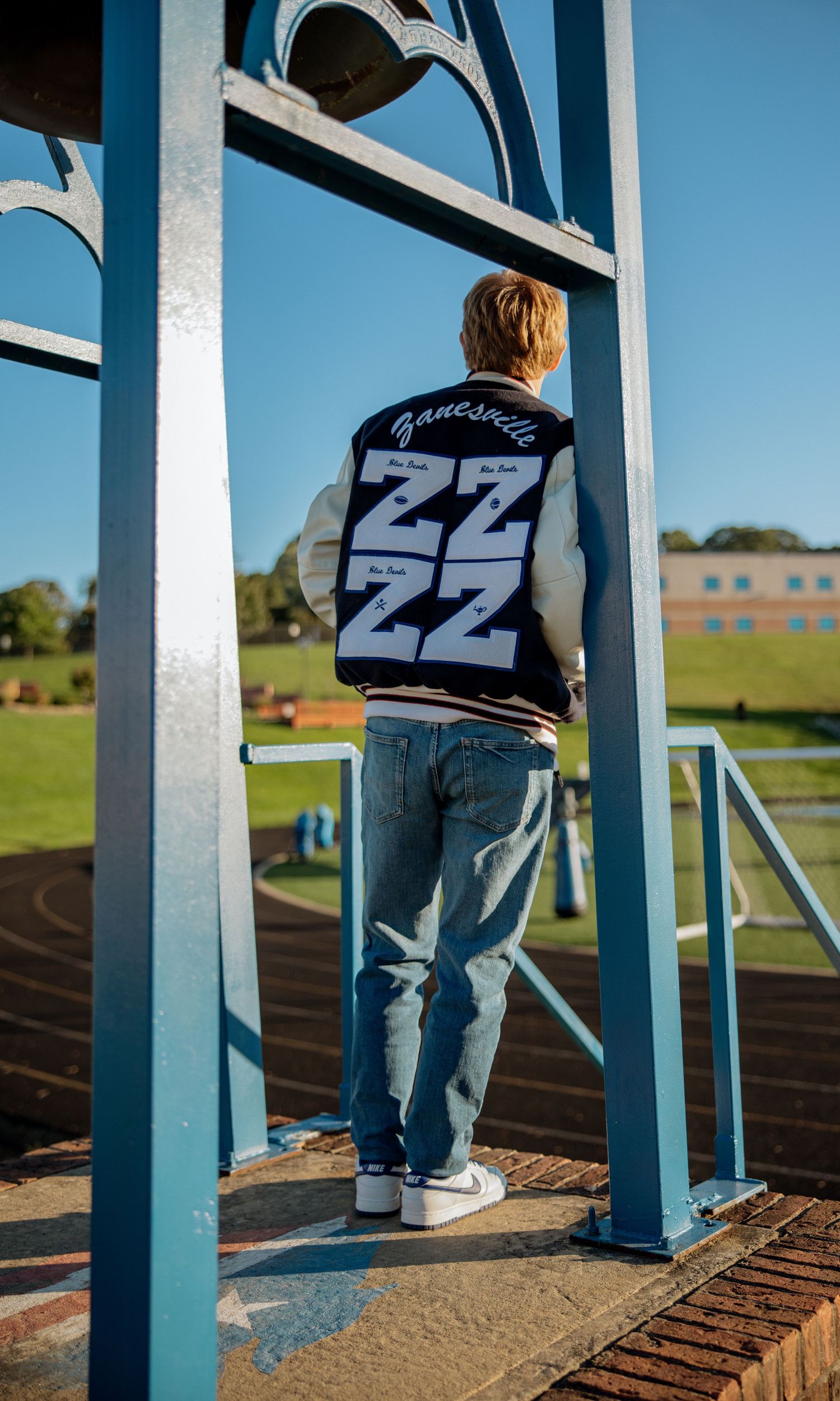 Person wearing a Danceville varsity jacket with ZZ lettering, standing on blue bleachers at a track field.