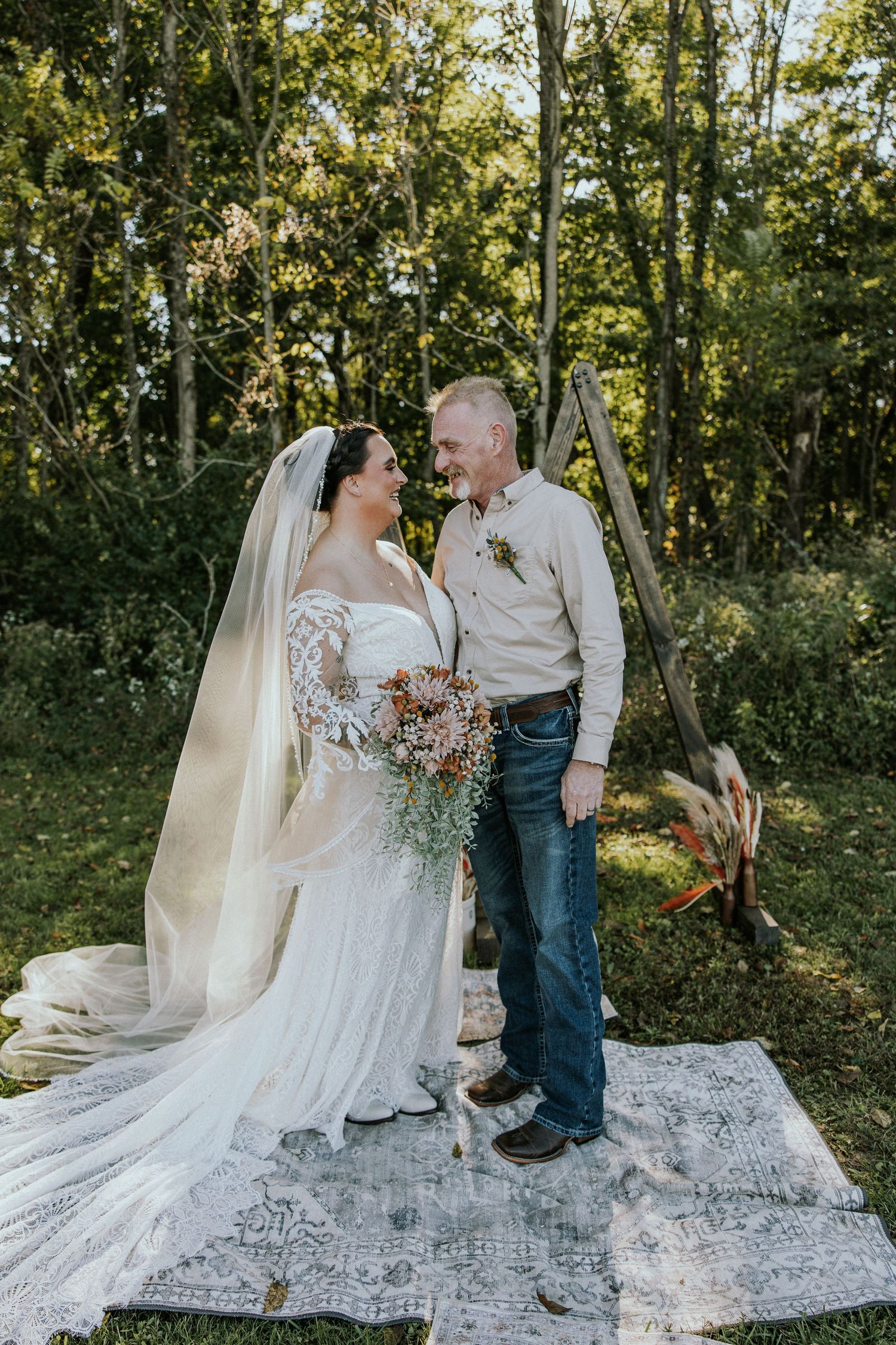 Bride and groom share a loving glance outdoors, standing on a vintage rug before a wooden arch surrounded by lush greenery.