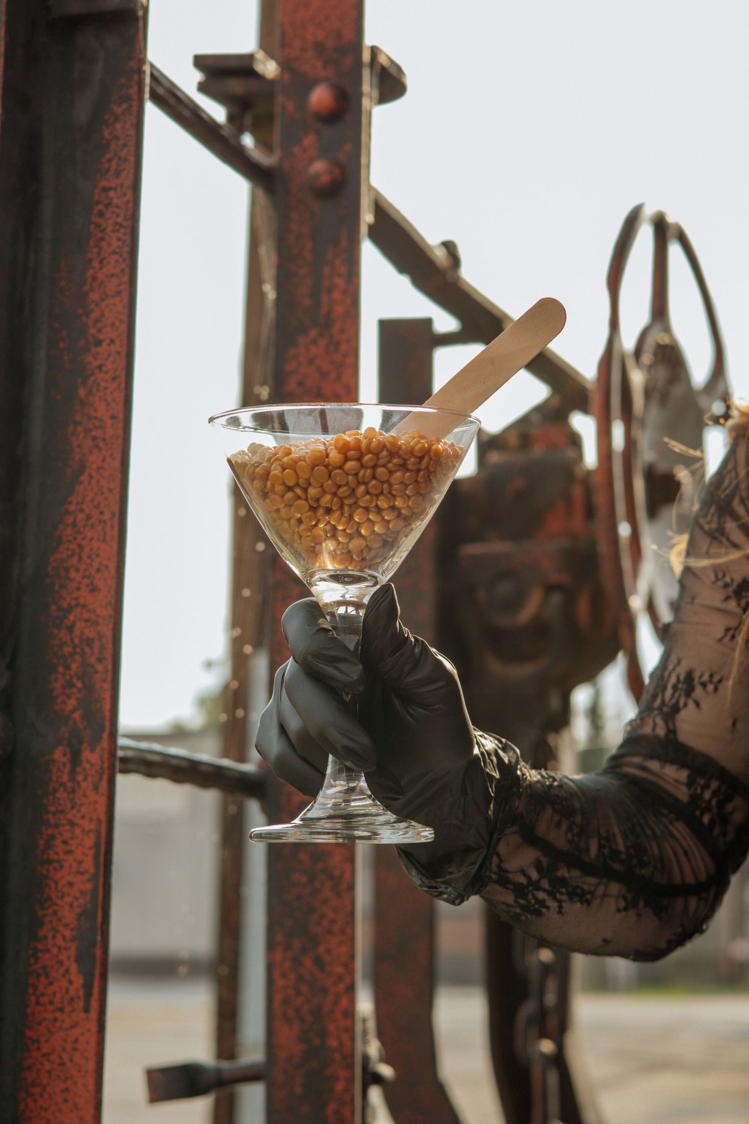 Gloved hand holding martini glass filled with golden wax beads with wooden stick near rusty metal structure.