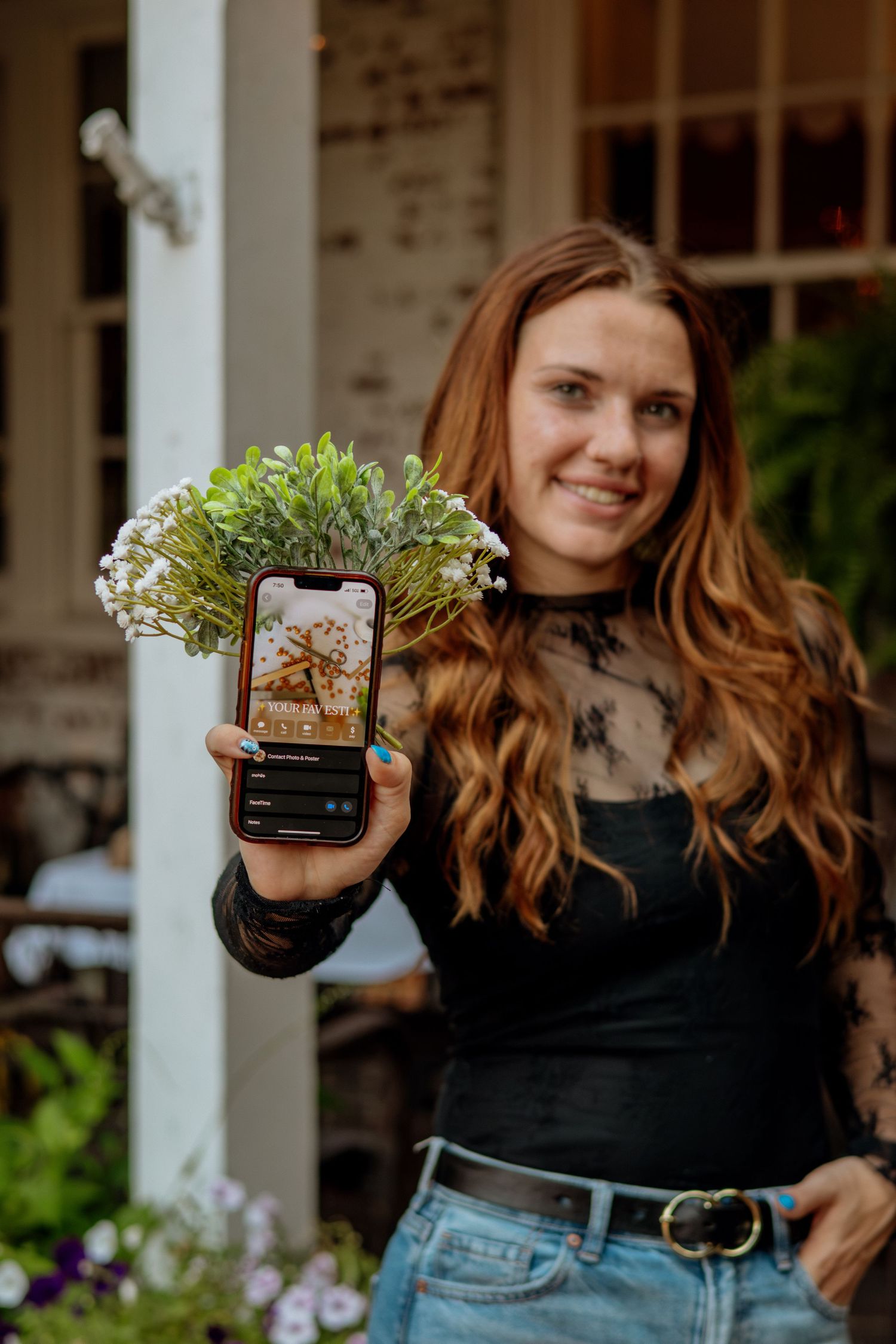 Smiling woman holding phone and flowers outdoors, displaying social media content on screen near porch.