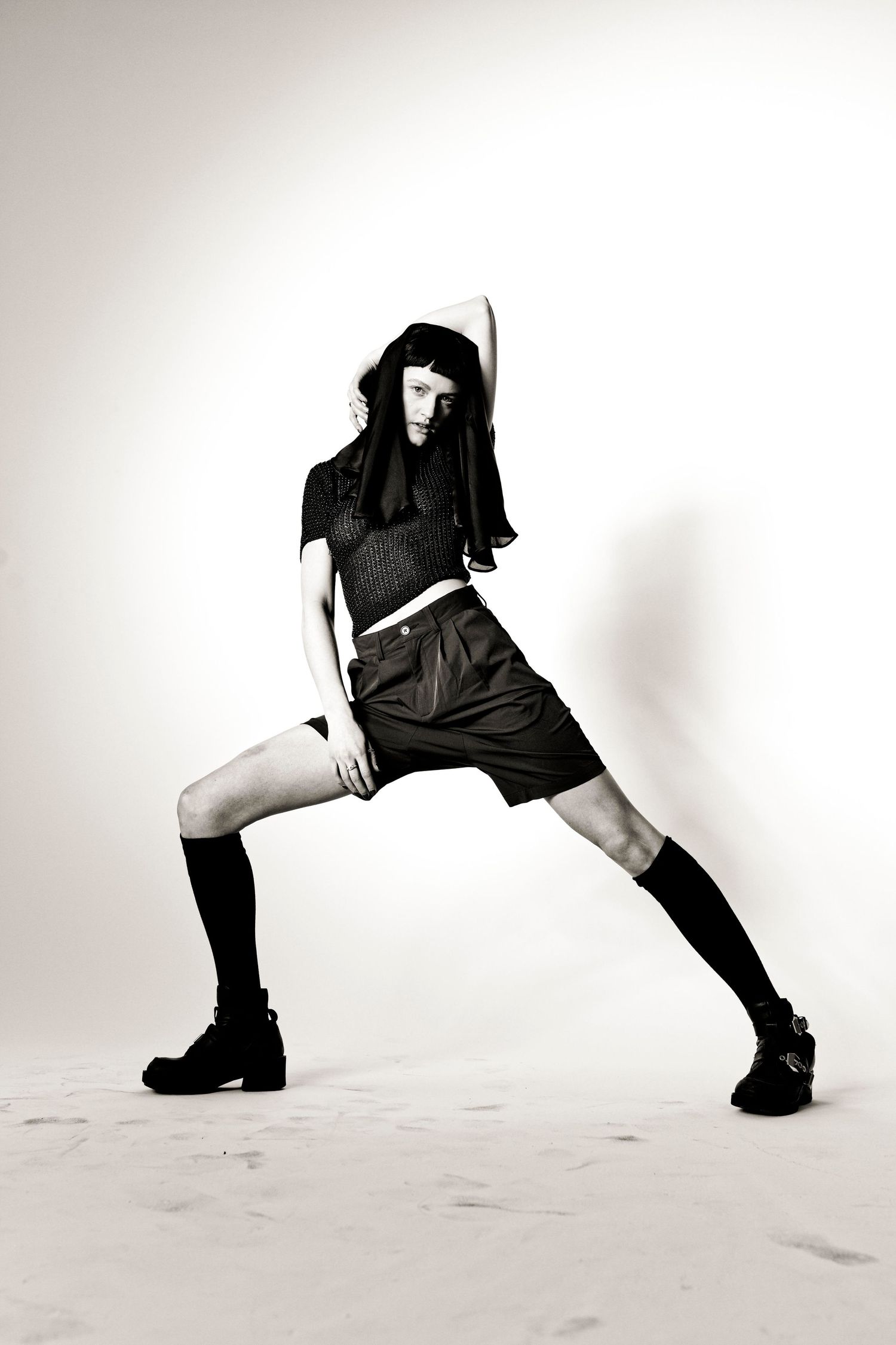 Black and white fashion photo of a model in knee socks, shorts, and crop top striking a wide-legged pose.