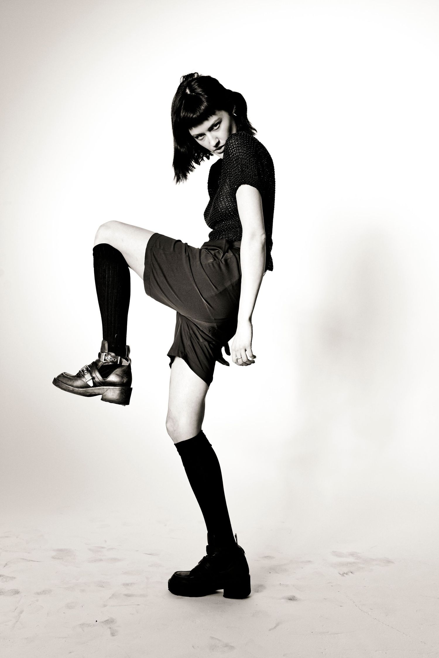 Black and white photo of a young woman in knee-high socks and platform shoes striking a dynamic pose.