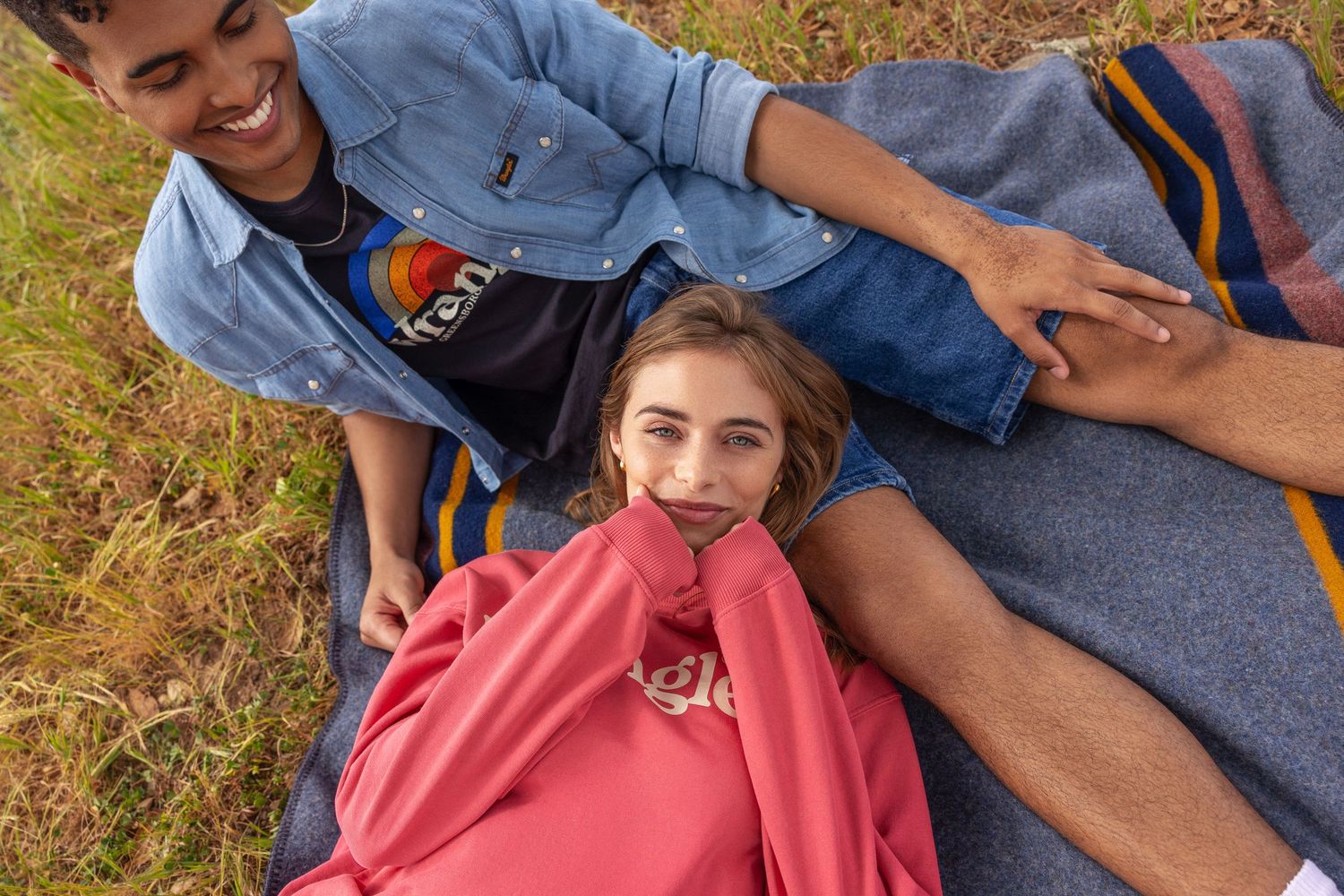 Young couple relaxing on blanket outdoors, girl lying down wrapped in pink sweatshirt smiling.