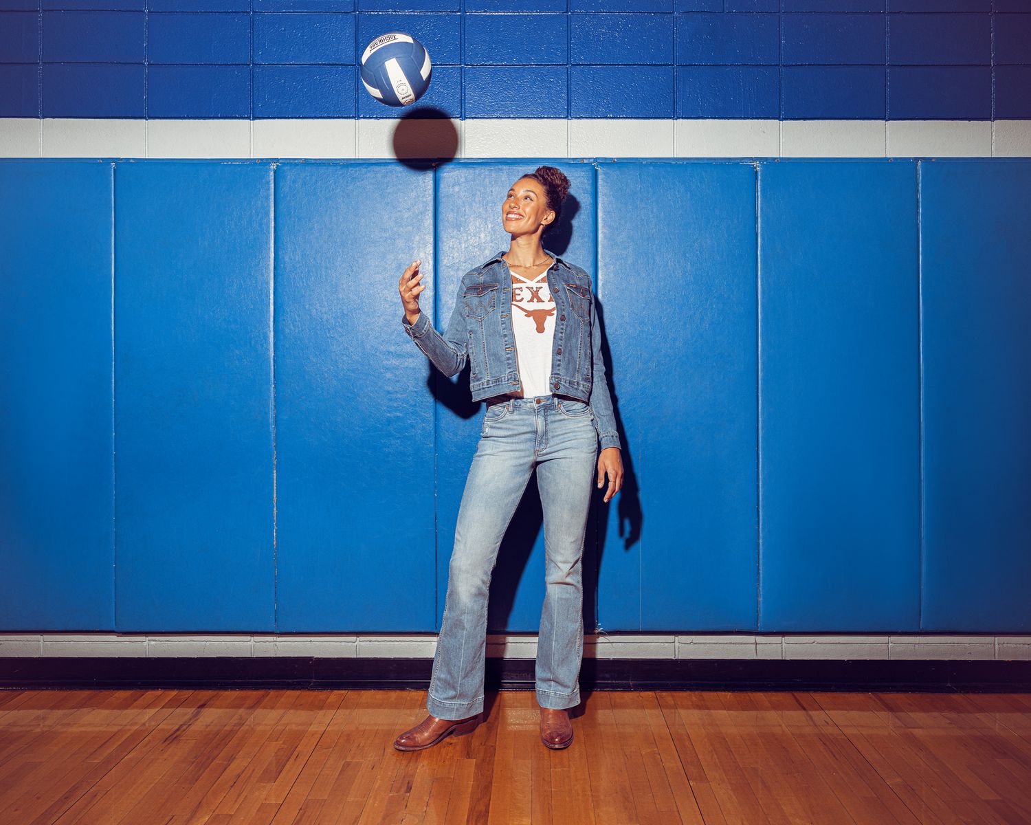 Woman in jeans and denim jacket tossing a volleyball in a gym with blue walls.