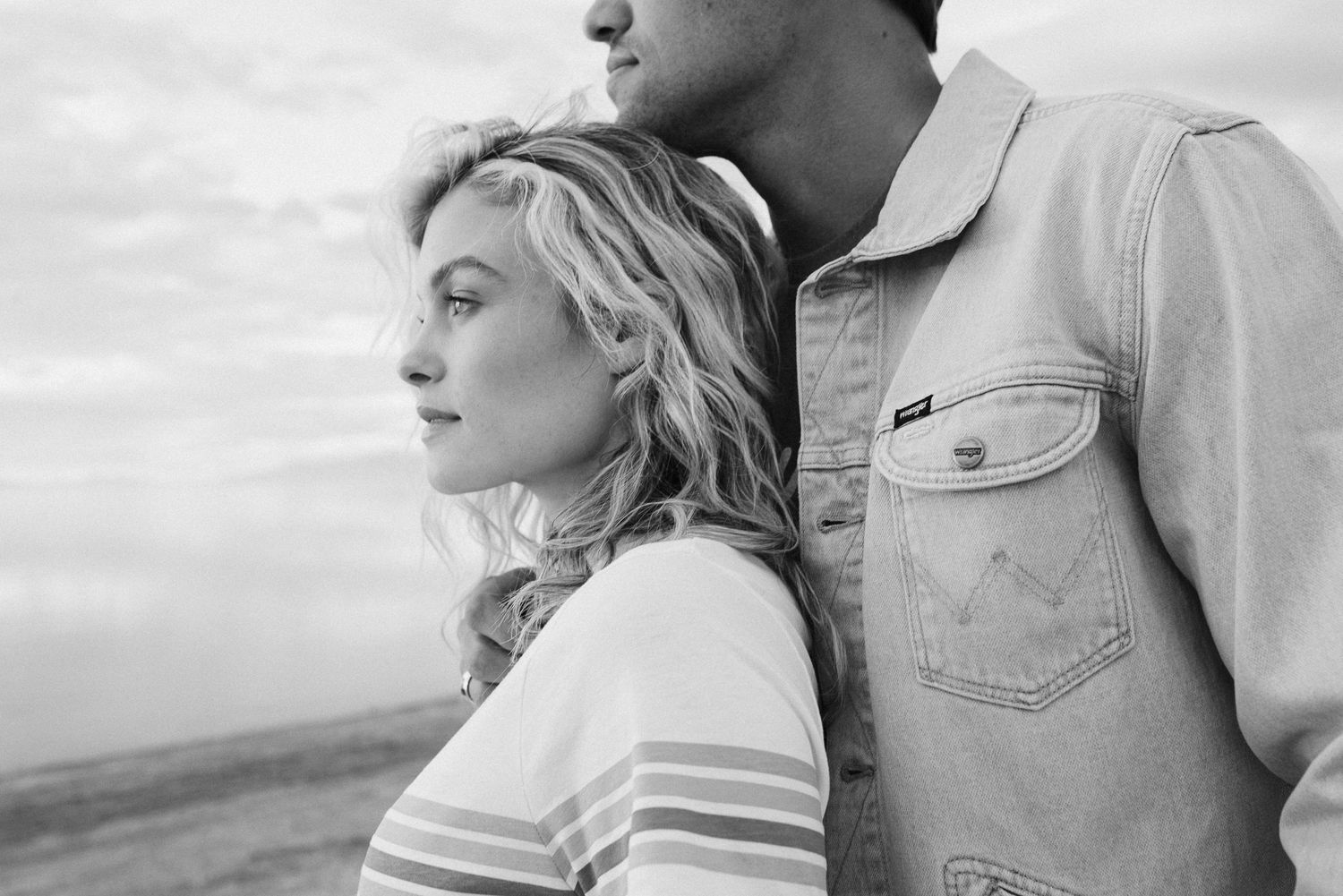 Black and white photo of a blonde woman and man standing together against a cloudy sky.