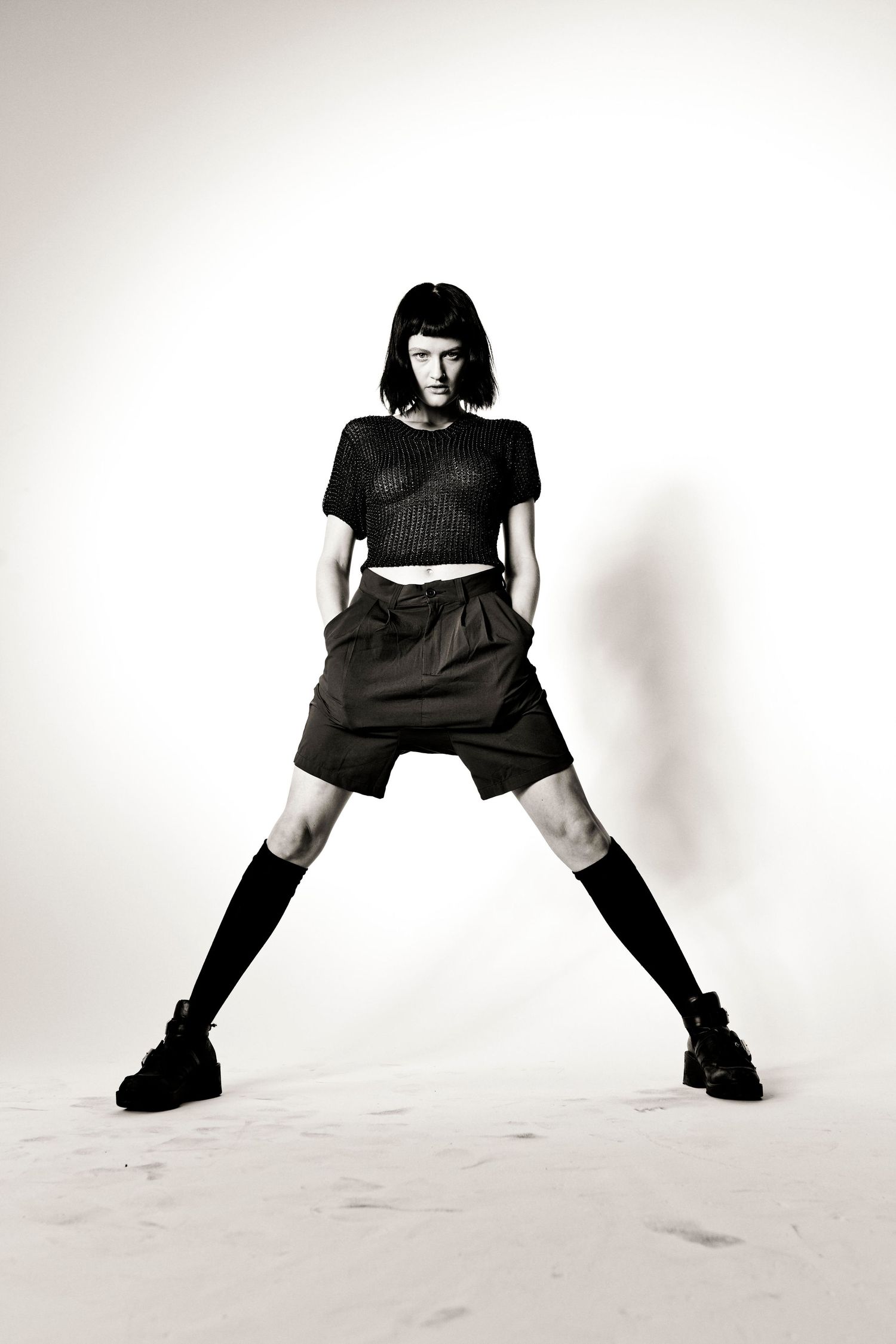 Black and white fashion photo of a model in crop top, shorts, and knee-high socks posing with legs apart.