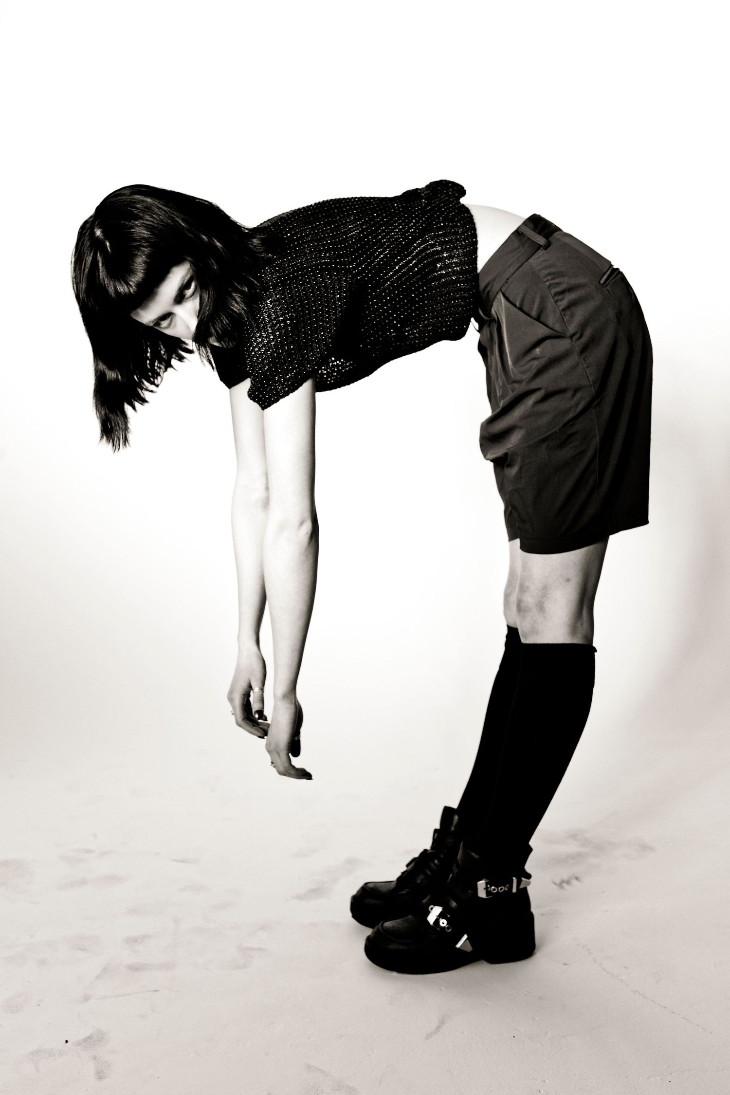 Black and white photo of a person bending forward in black mesh top, shorts, knee-high socks, and buckled boots.