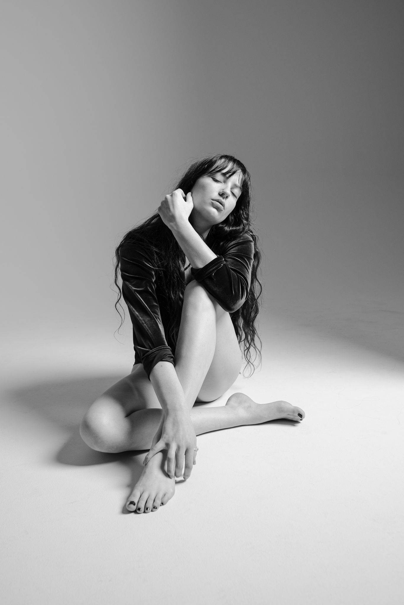 Black and white photo of a brunette woman in a dark jacket sitting barefoot on a studio floor.