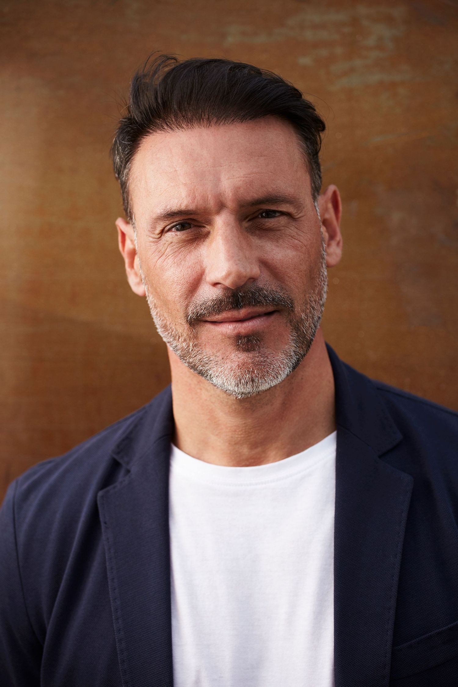 Middle-aged man with salt-and-pepper stubble wearing a navy blazer and white t-shirt posing against a brown background.