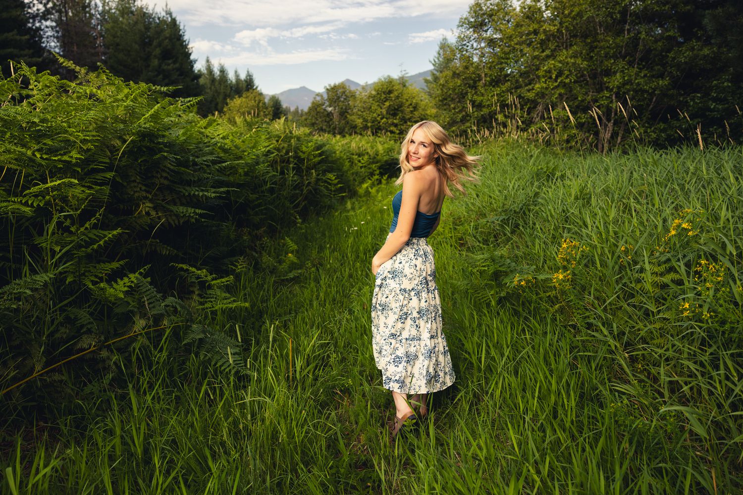 Joyful blonde woman in floral skirt spinning on green nature path surrounded by lush ferns and foliage.