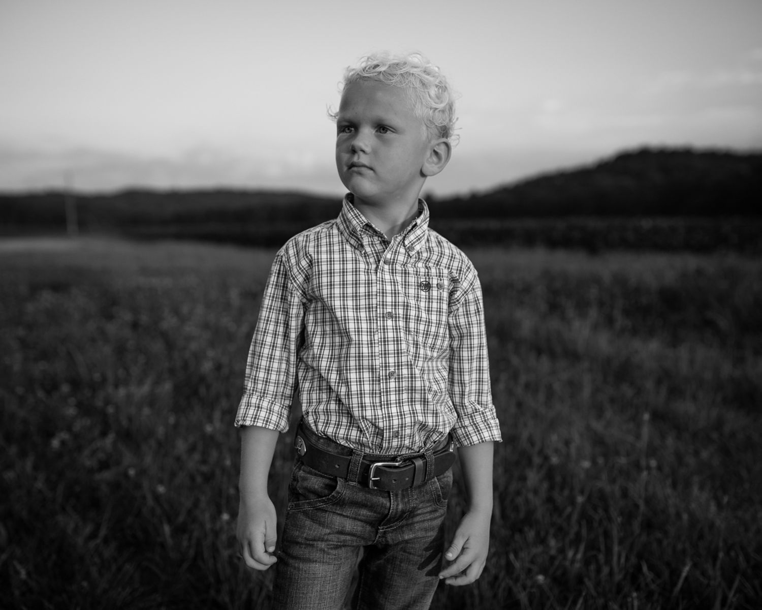 Blonde boy in plaid shirt and jeans standing in open field at dusk, black and white portrait.
