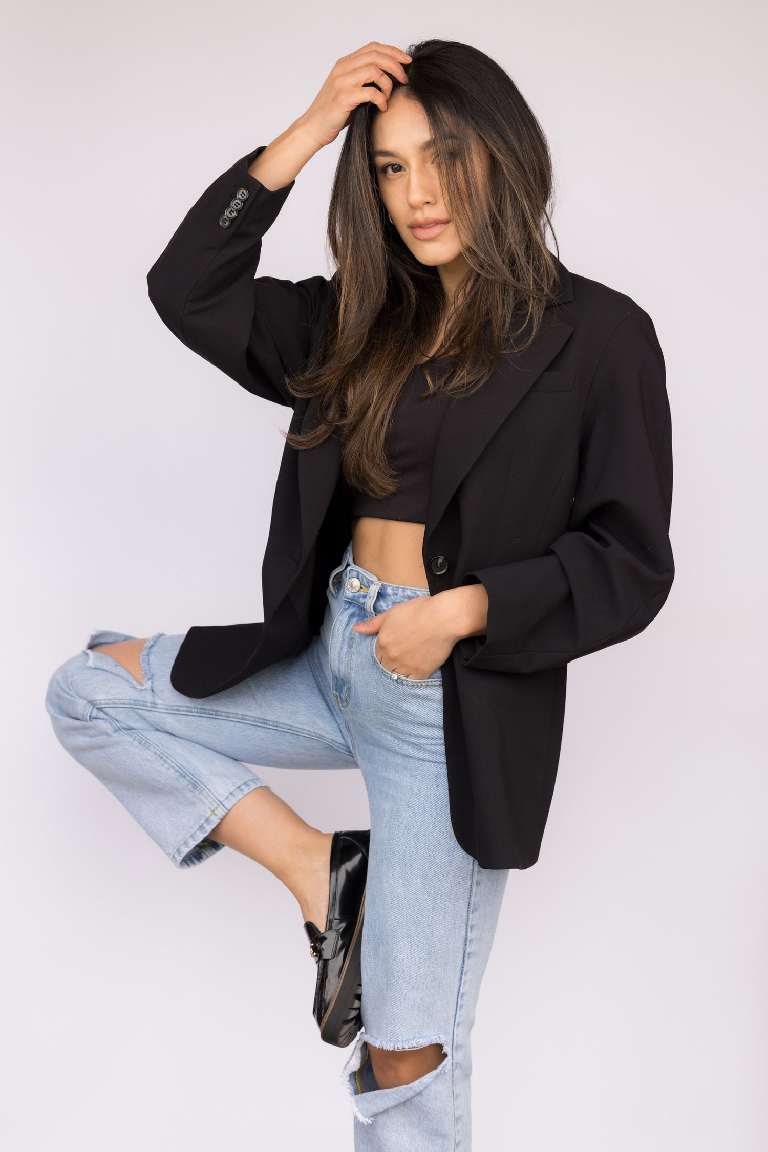 Woman in black blazer, crop top, and ripped jeans posing in studio.