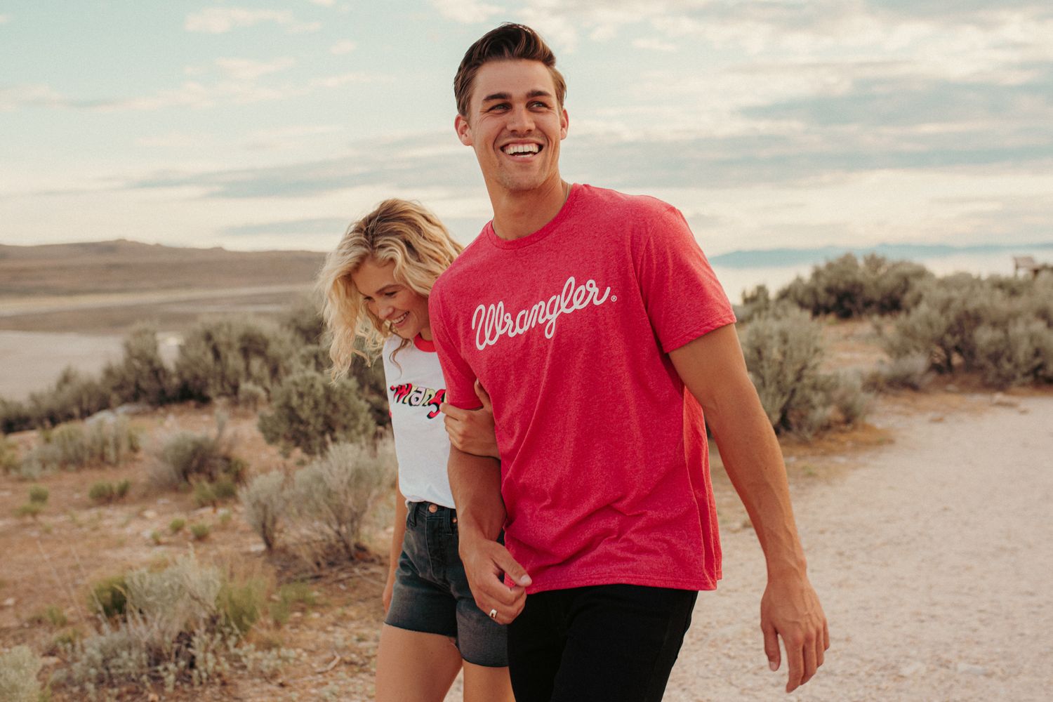 A smiling couple walking hand-in-hand outdoors wearing Wrangler branded t-shirts.