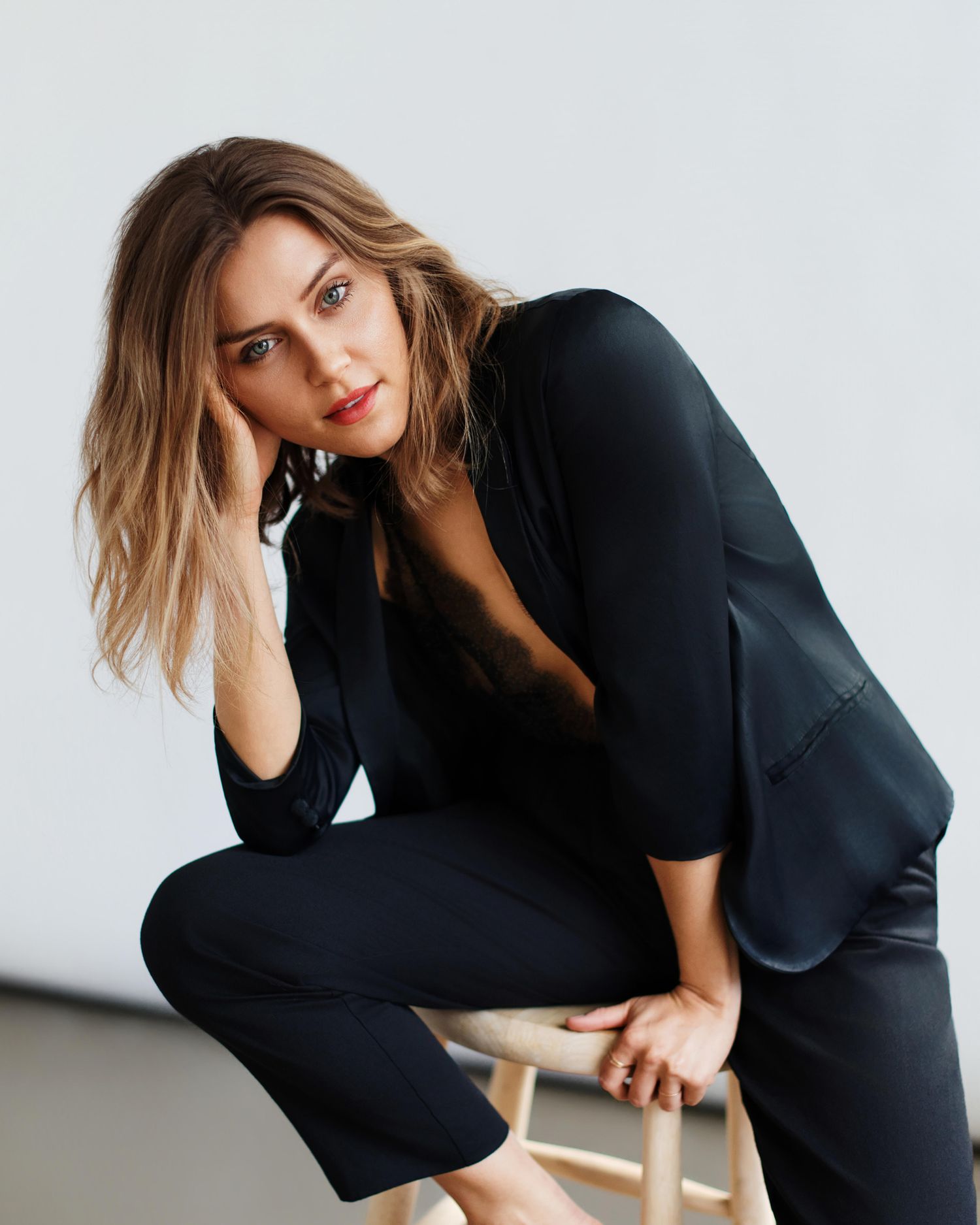 Woman in black outfit sitting on stool, posing with hand on cheek, studio portrait photography.