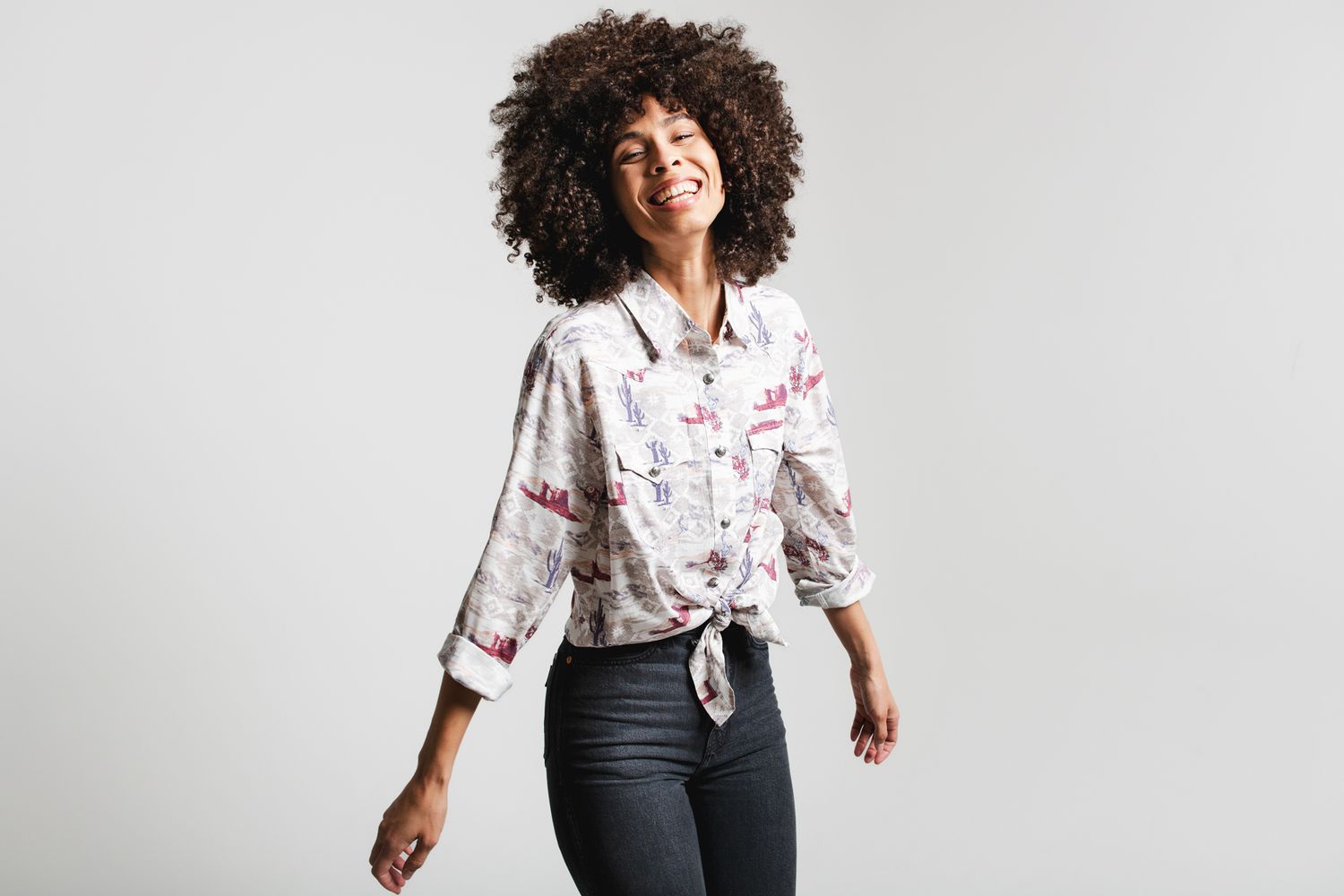 Cheerful woman with curly hair wearing a floral tie-front shirt and dark jeans on gray background.