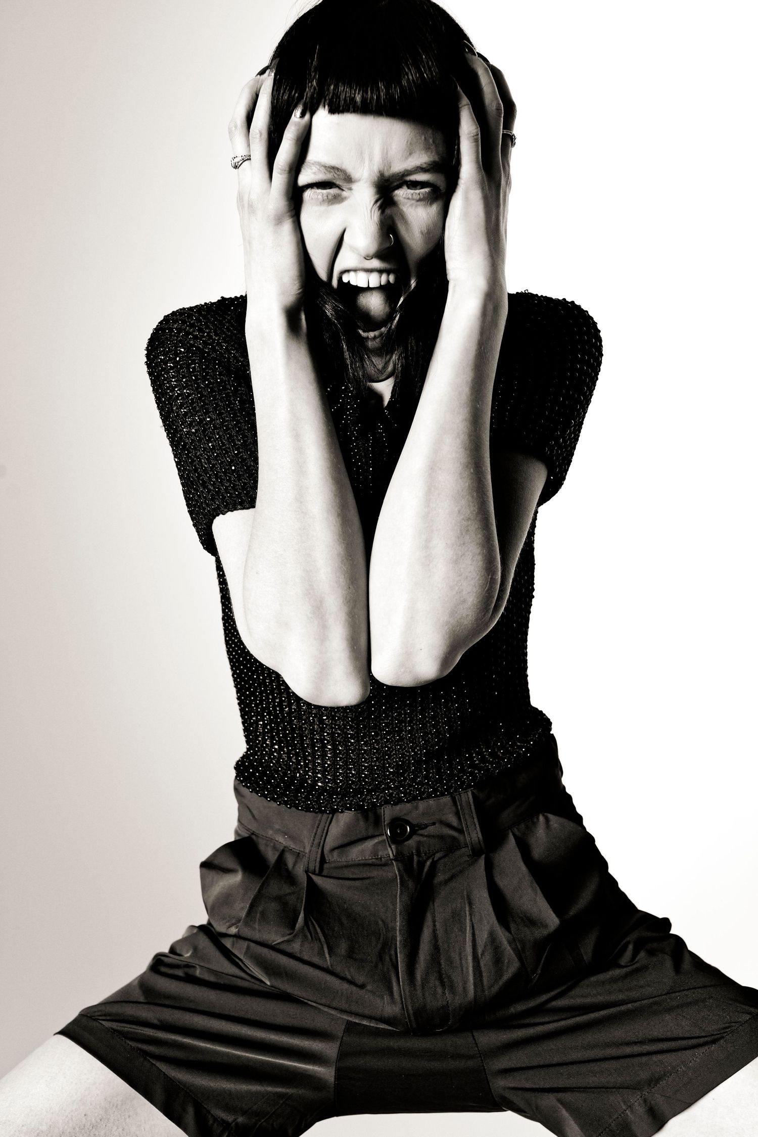 Black and white photo of a woman screaming with hands on her face, wearing a black top and wide-leg pants.