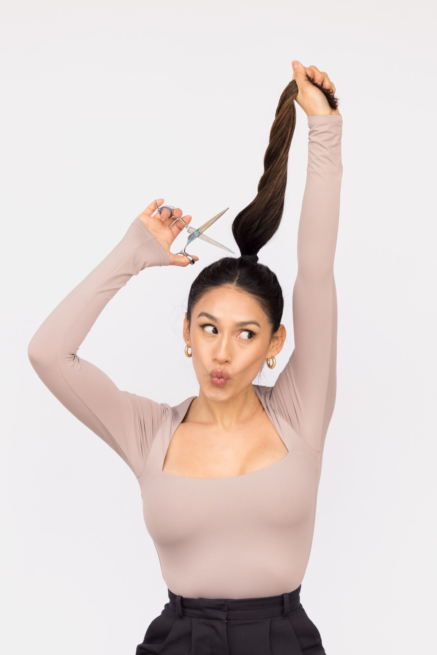 Young woman playfully holding her ponytail up with scissors as if cutting her long dark hair, wearing a beige bodysuit.