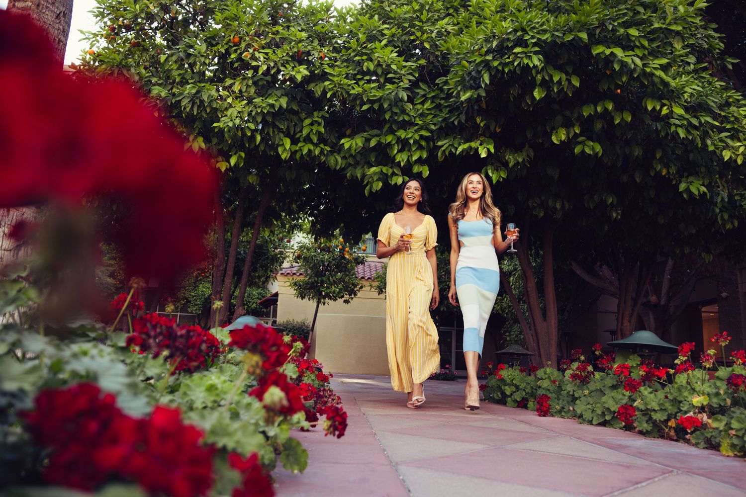 Two women in colorful summer dresses walking through a lush garden with red roses.
