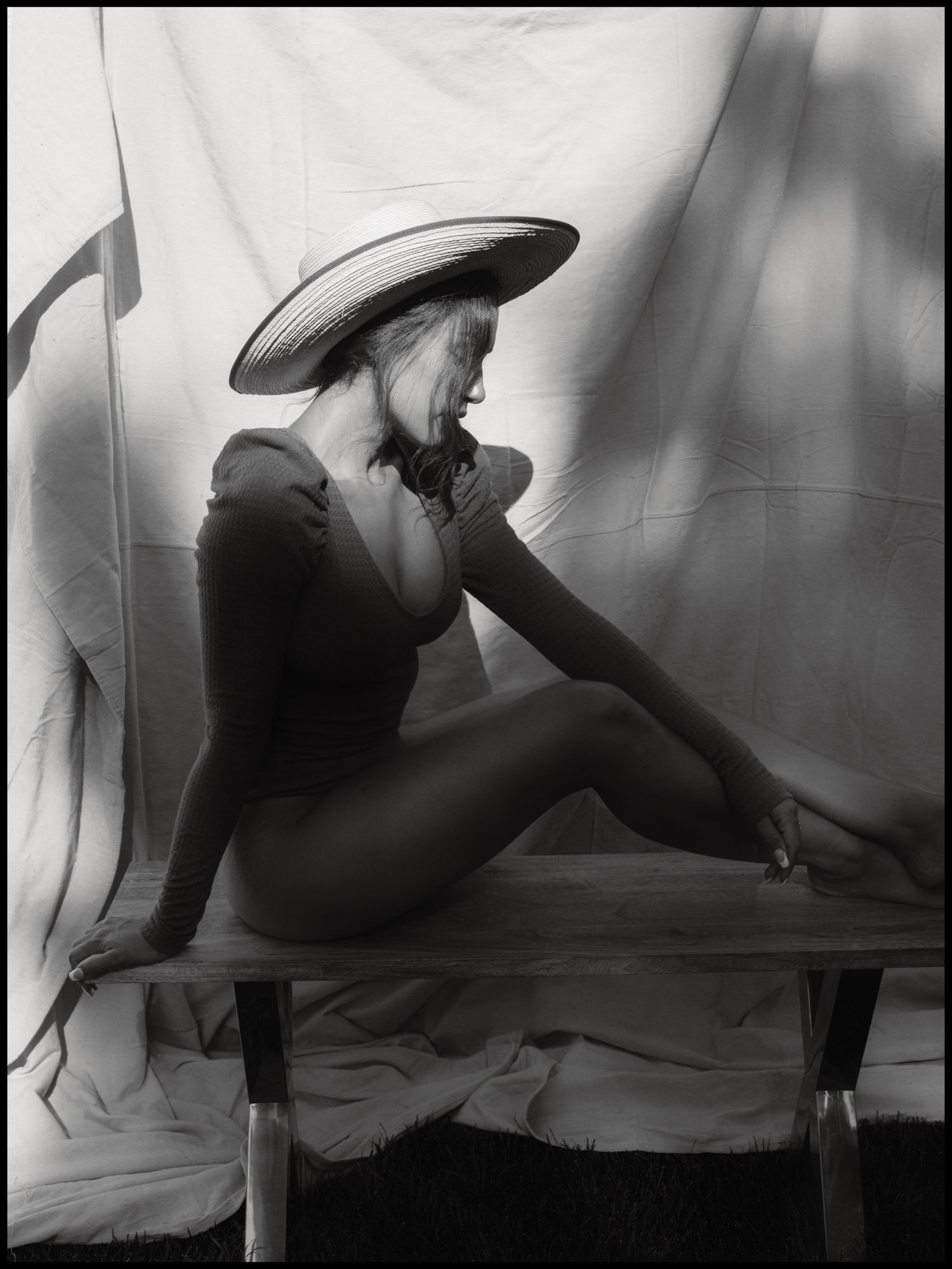 Elegant woman in wide-brim hat sitting on bench in graceful pose, artistic black and white photography.