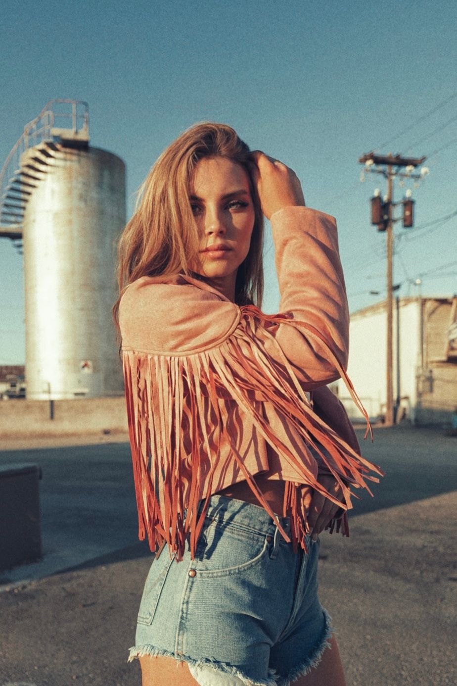 Young woman in a pink fringe suede jacket and denim shorts posing outdoors near industrial silos at golden hour.
