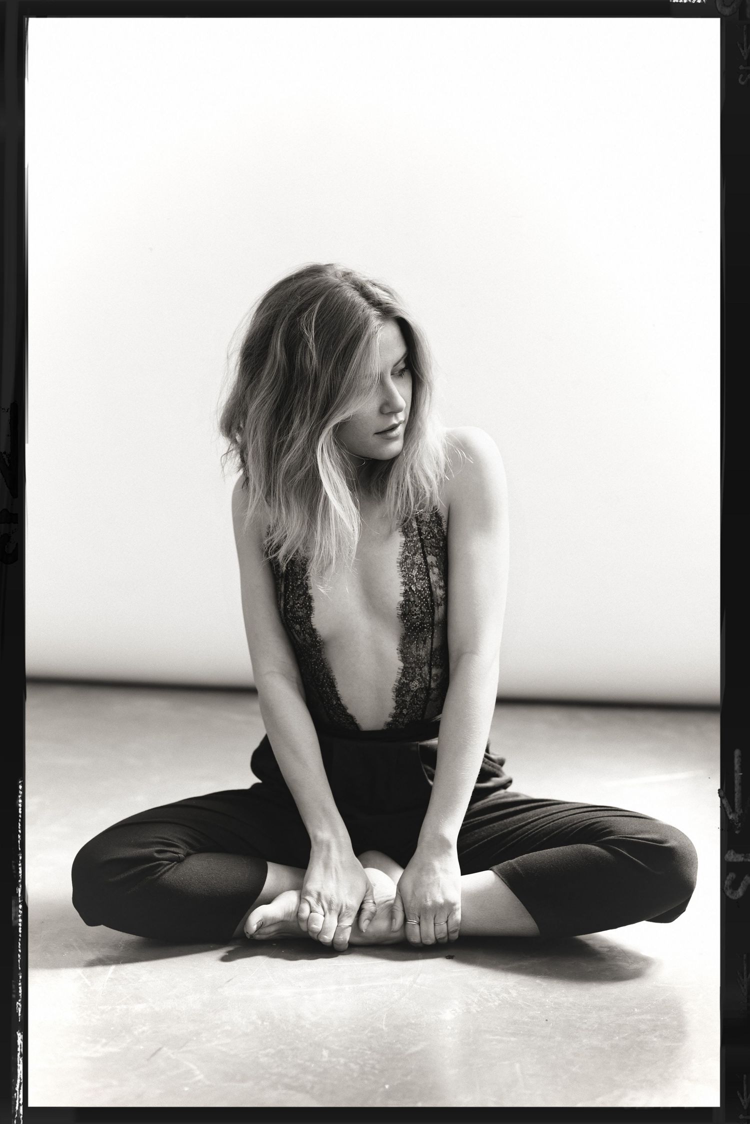 Black and white photo of a woman sitting cross-legged on the floor in a lace bodysuit and dark trousers, head bowed.