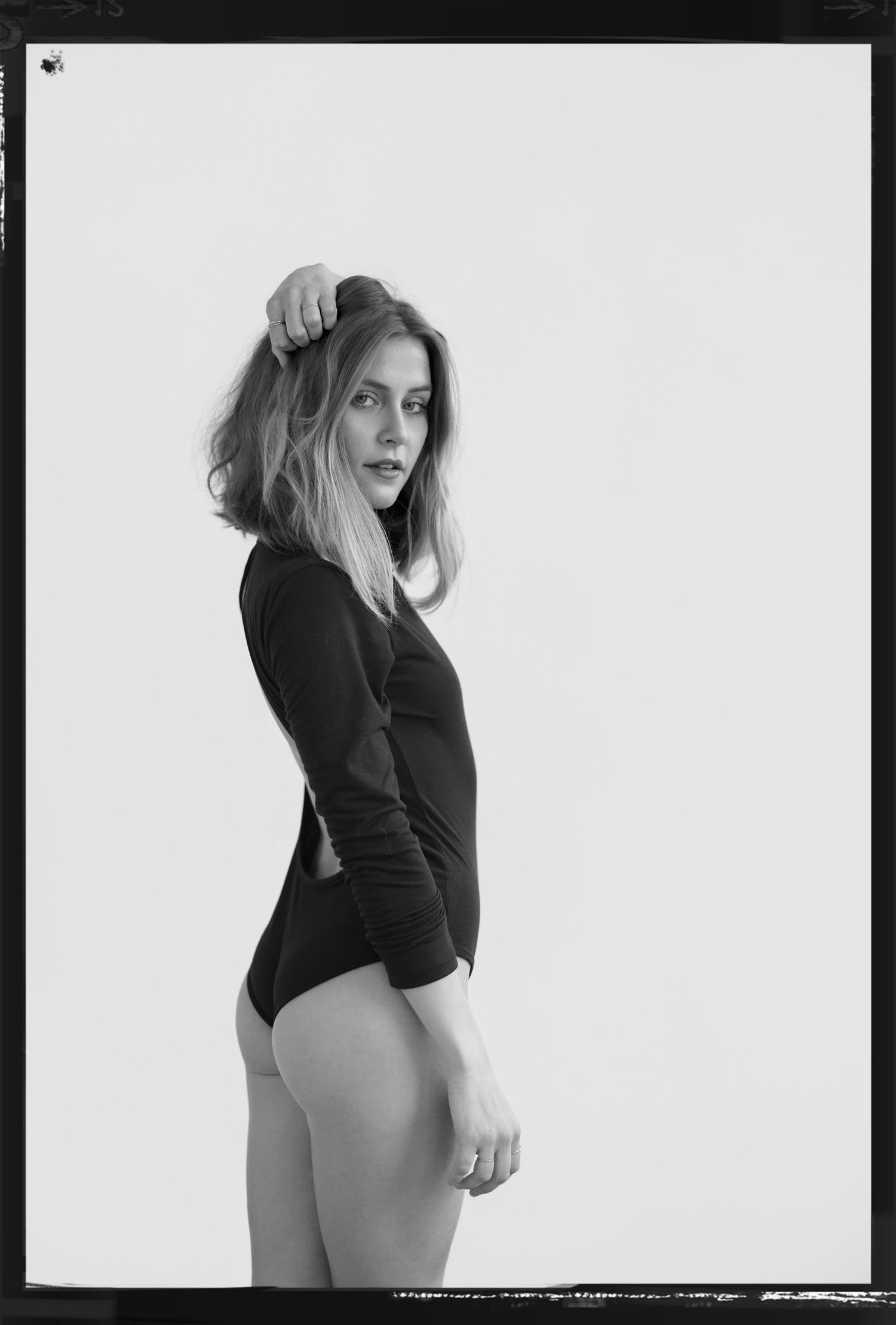 Black and white studio photo of a woman in a black long-sleeve bodysuit posing sideways against a white background.