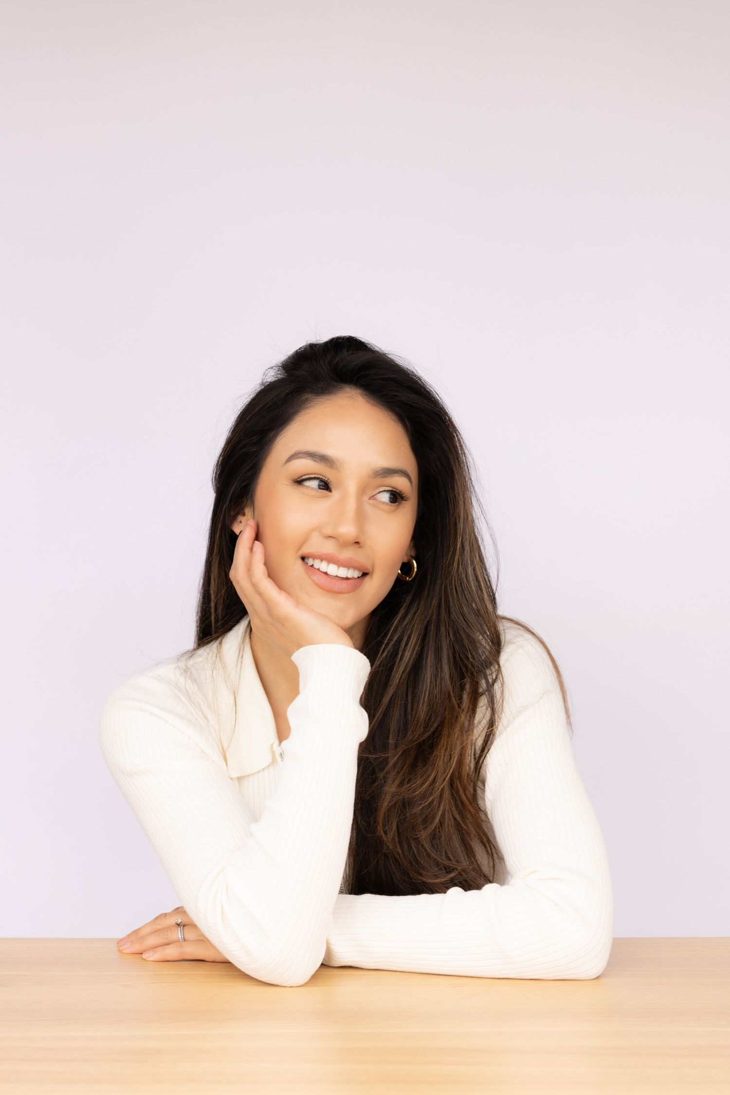 Smiling woman in white turtleneck resting chin on hand at wooden table.