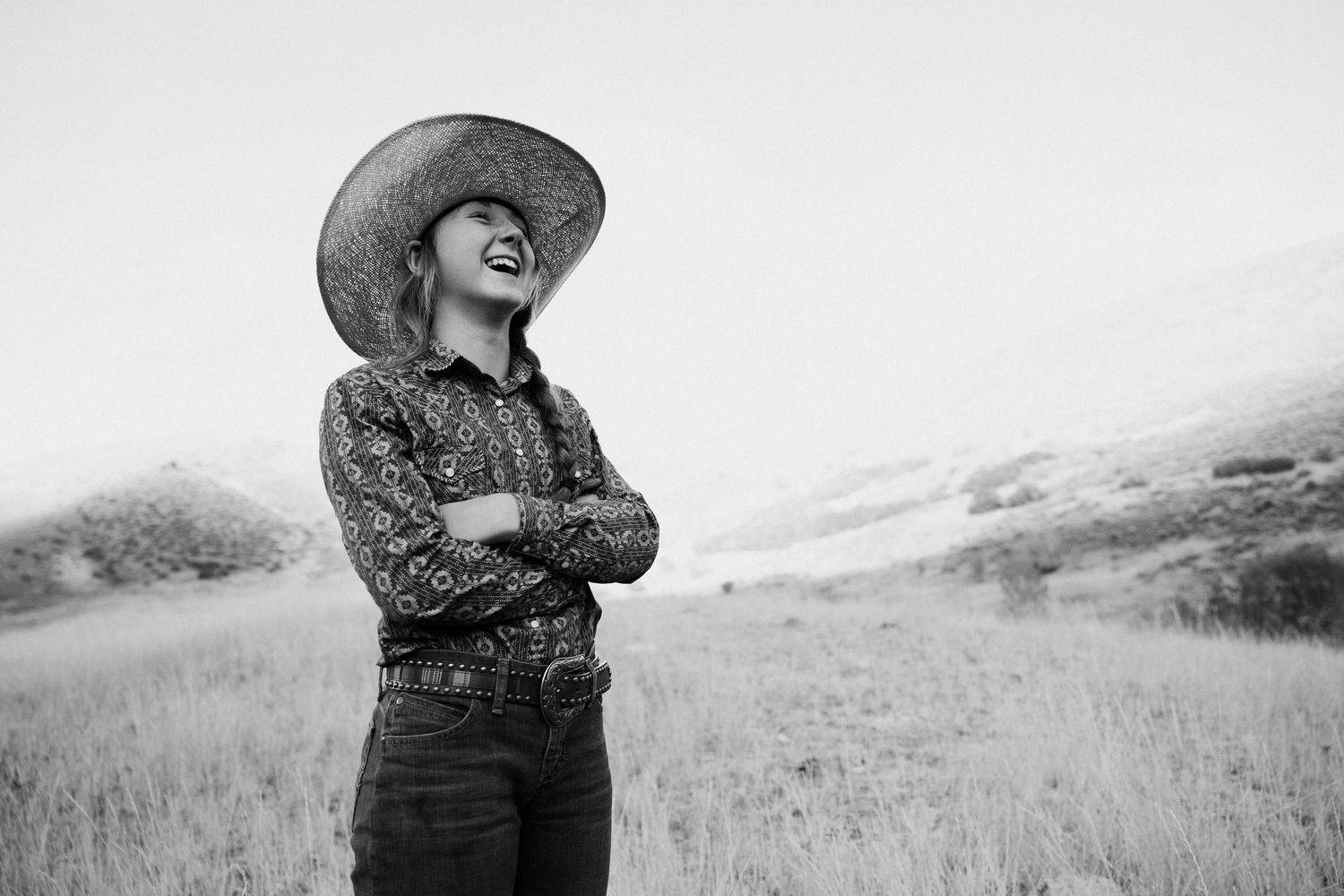 Black and white photo of a laughing woman in a wide-brim hat with arms crossed standing in an open field.