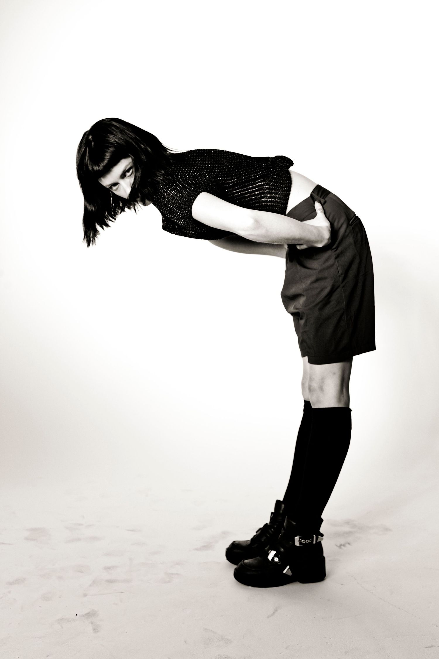 Black and white photo of a woman in dark knit top, mini skirt, knee socks, and chunky shoes, bending forward.