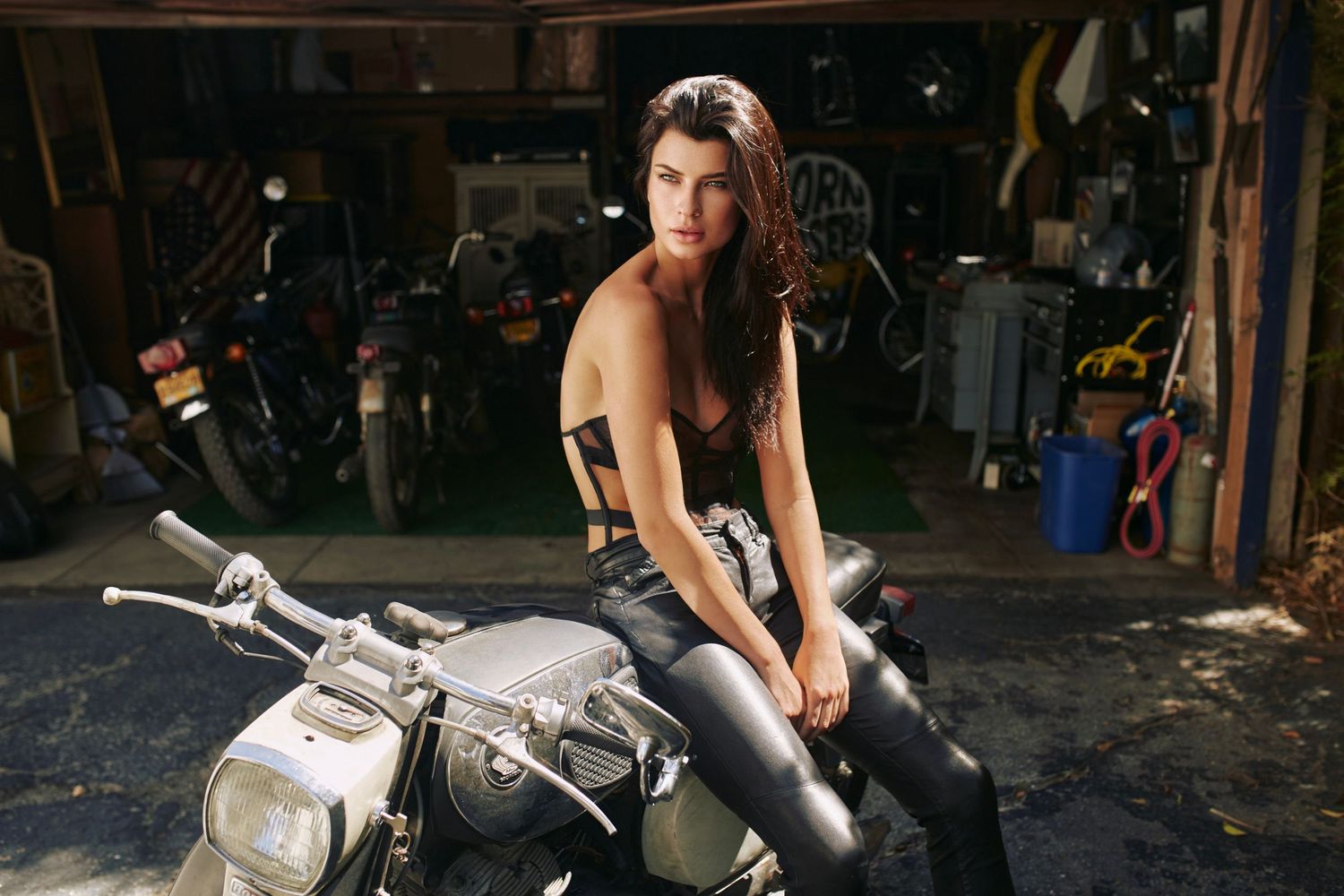 Woman in black outfit sitting on a vintage motorcycle inside a rustic garage workshop.