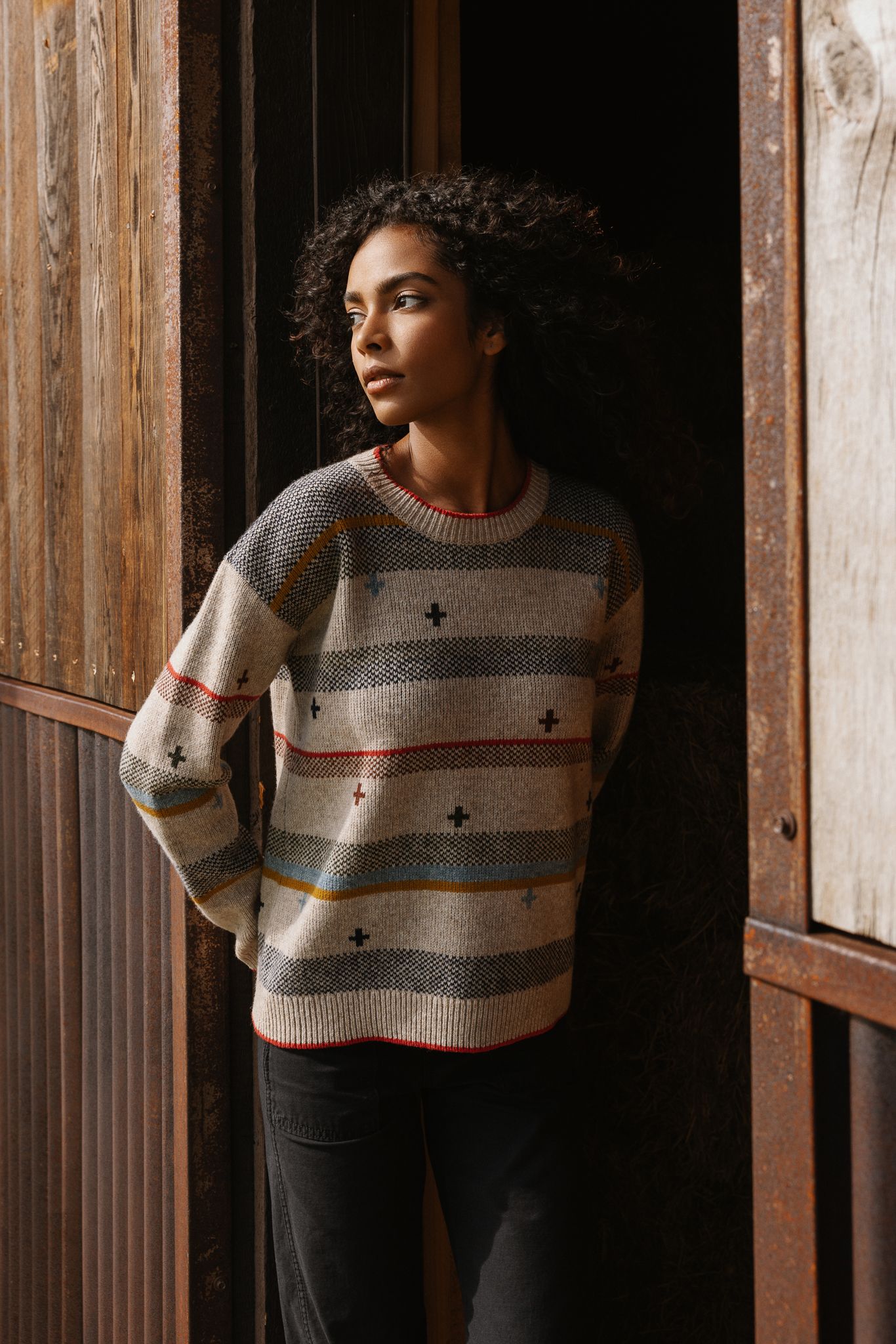 Woman with curly hair wearing patterned knit sweater leaning against rustic wooden wall, looking away.