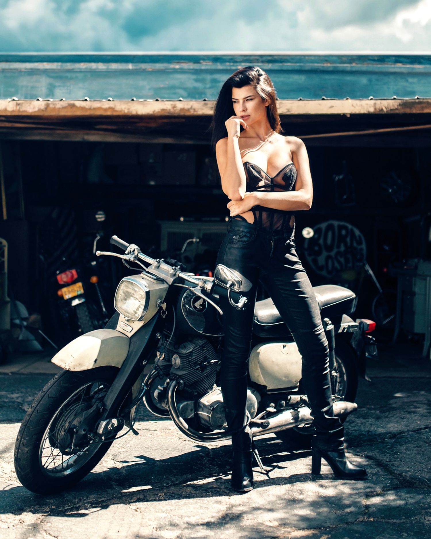 Woman in black leather outfit posing with a vintage motorcycle outside a garage on a cloudy day.