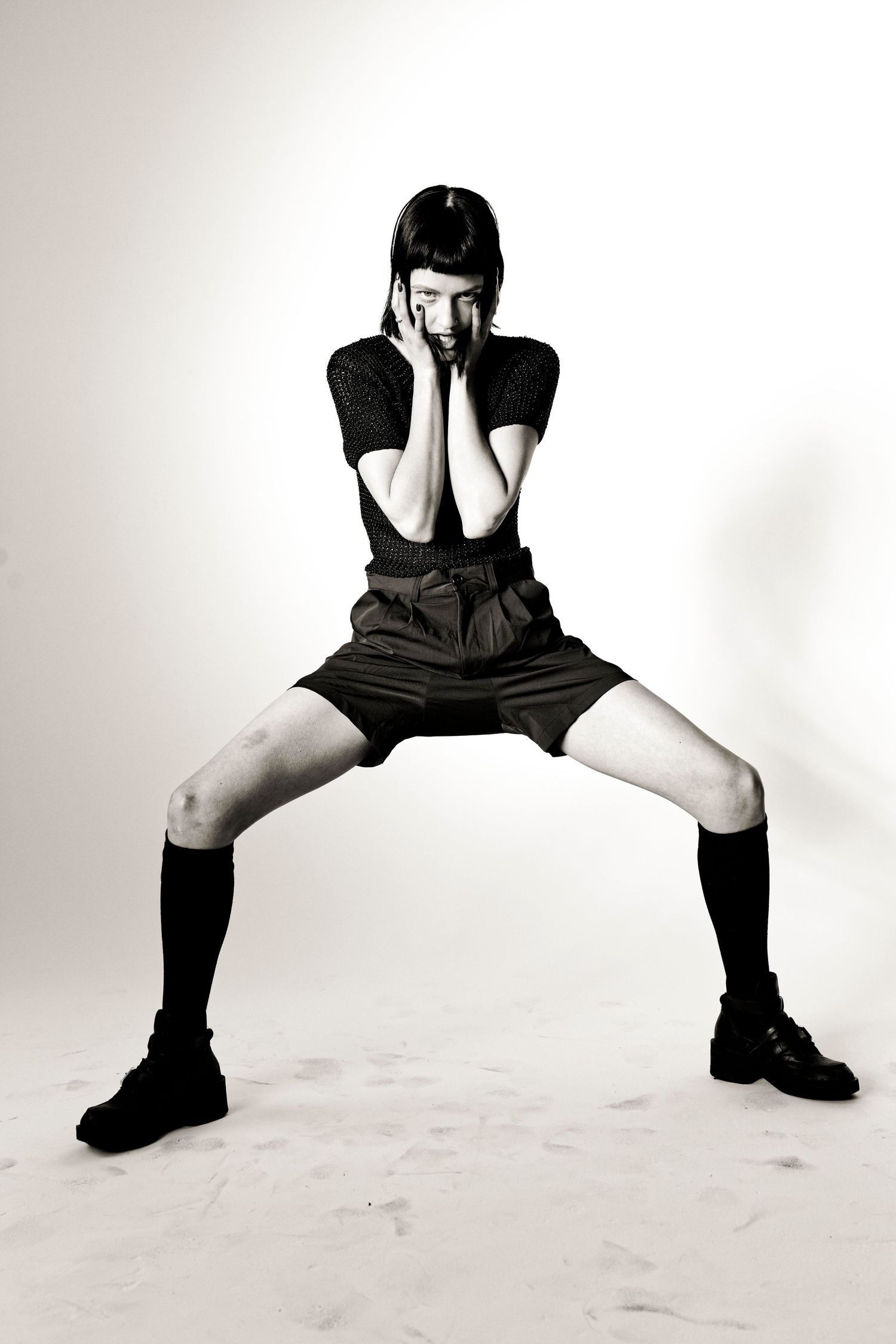Black and white photo of a young woman in shorts and knee socks posing with hands on face.
