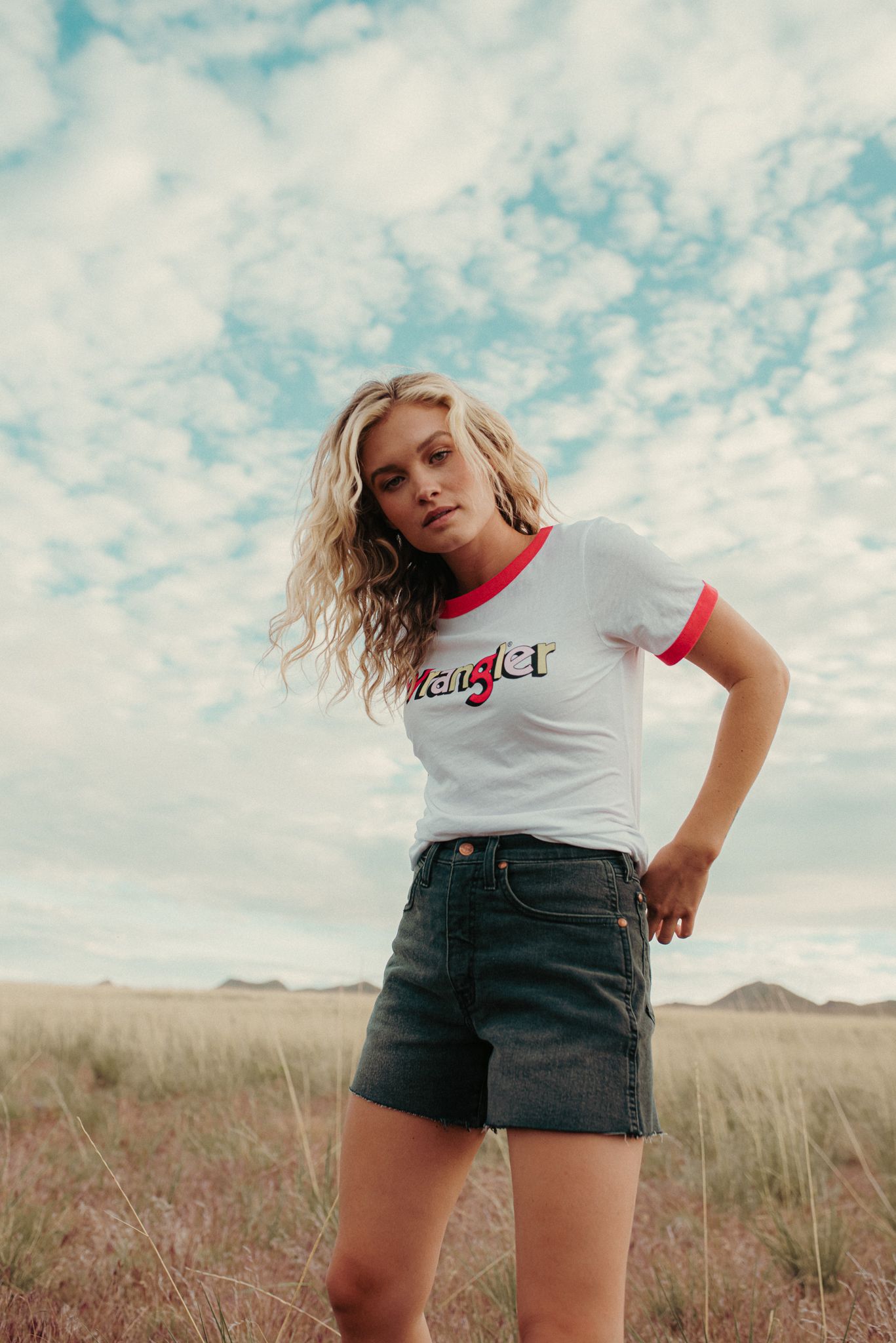 Blonde woman wearing a Wrangler ringer tee and denim shorts standing in an open field.