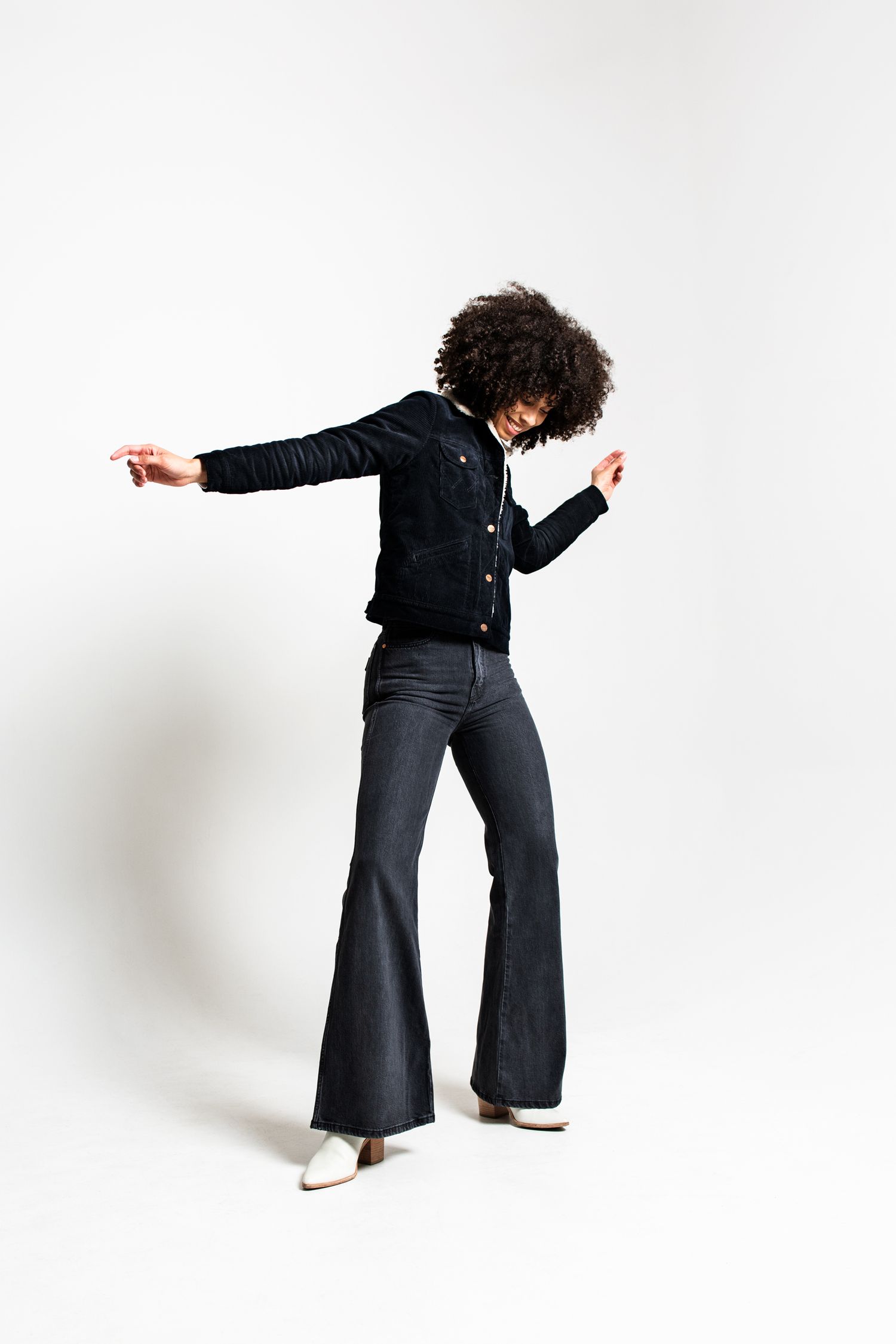 Full-length shot of a woman with curly hair dancing in flare jeans and black jacket on white background.