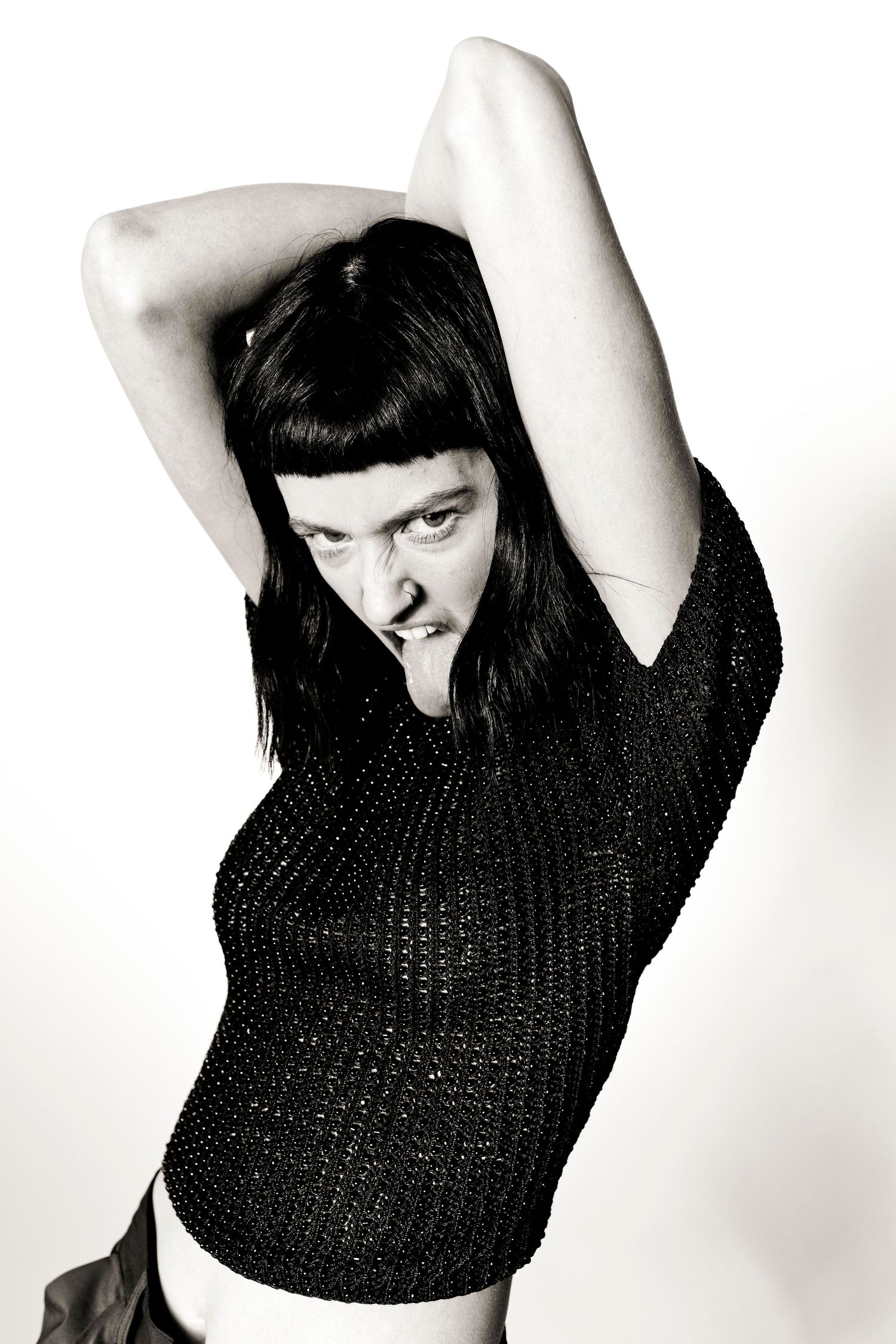 Woman in black mesh top poses dramatically with arms raised in black and white photo.