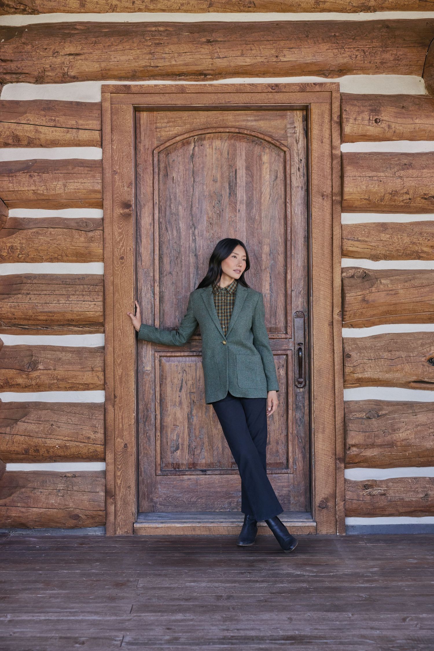 Woman in green blazer and black pants posing by rustic log cabin wooden door.