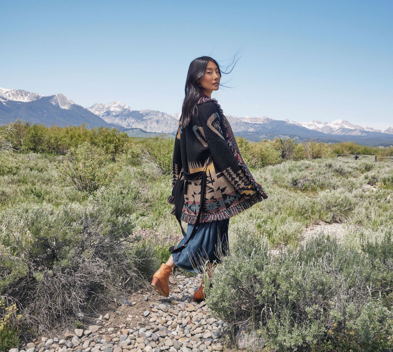 Woman in black patterned kimono walking through sage brush with mountain views.
