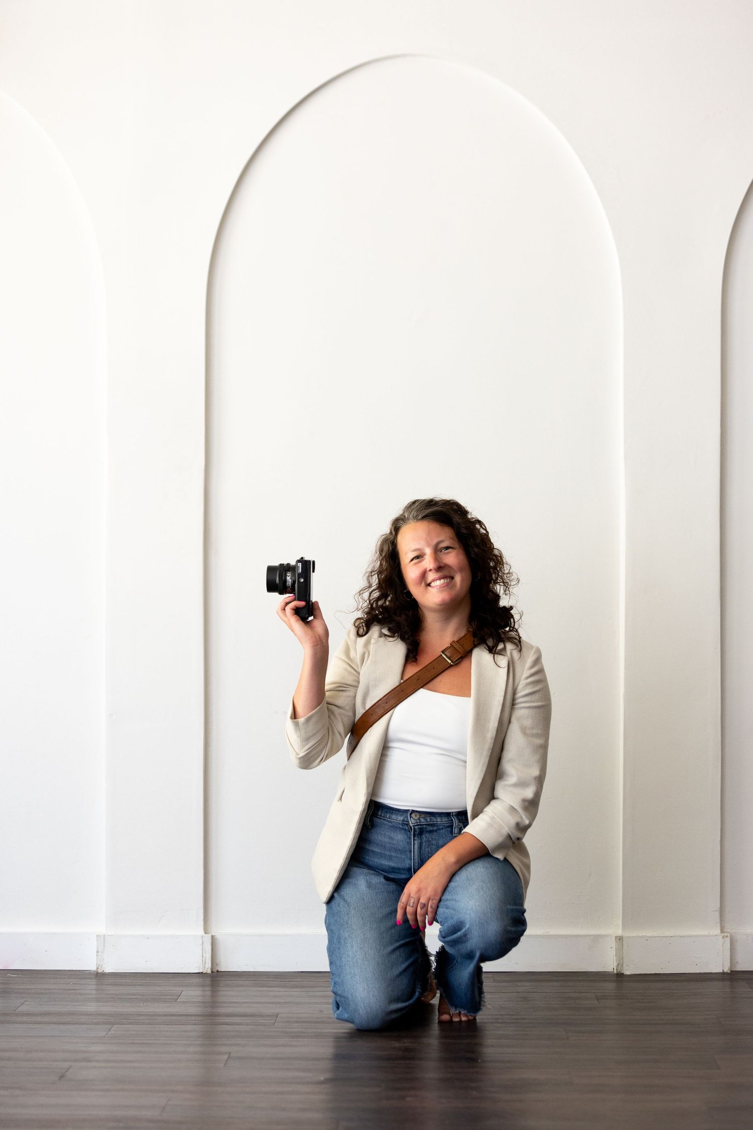 A smiling woman kneels on a dark wood floor holding a camera, wearing jeans and a blazer against a white arched wall.