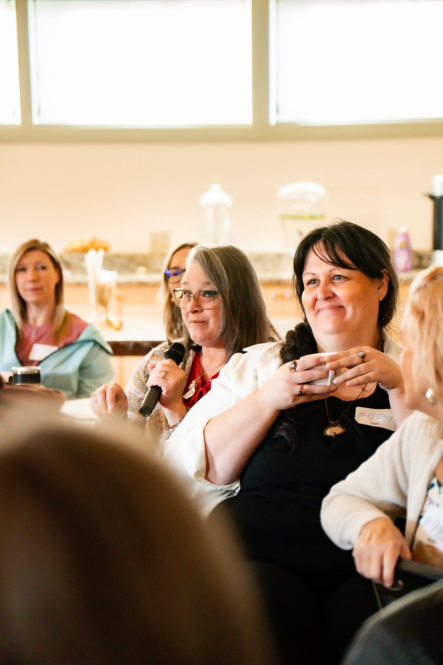Group of smiling women seated in a bright room, with one holding a microphone while others listen and laugh during a discussion event.