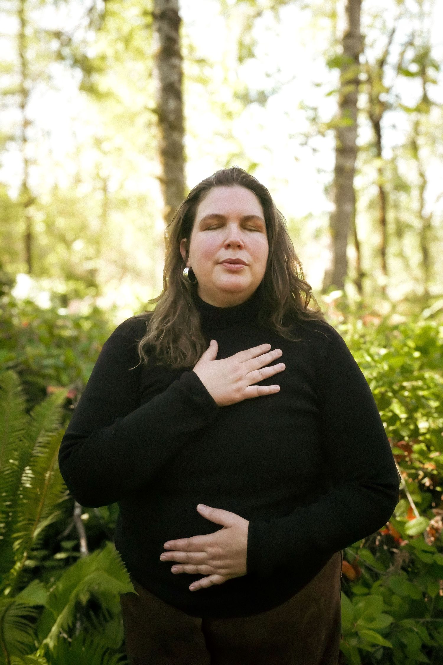 Woman in black turtleneck meditating outdoors in lush green forest, hands on chest and belly, eyes closed.