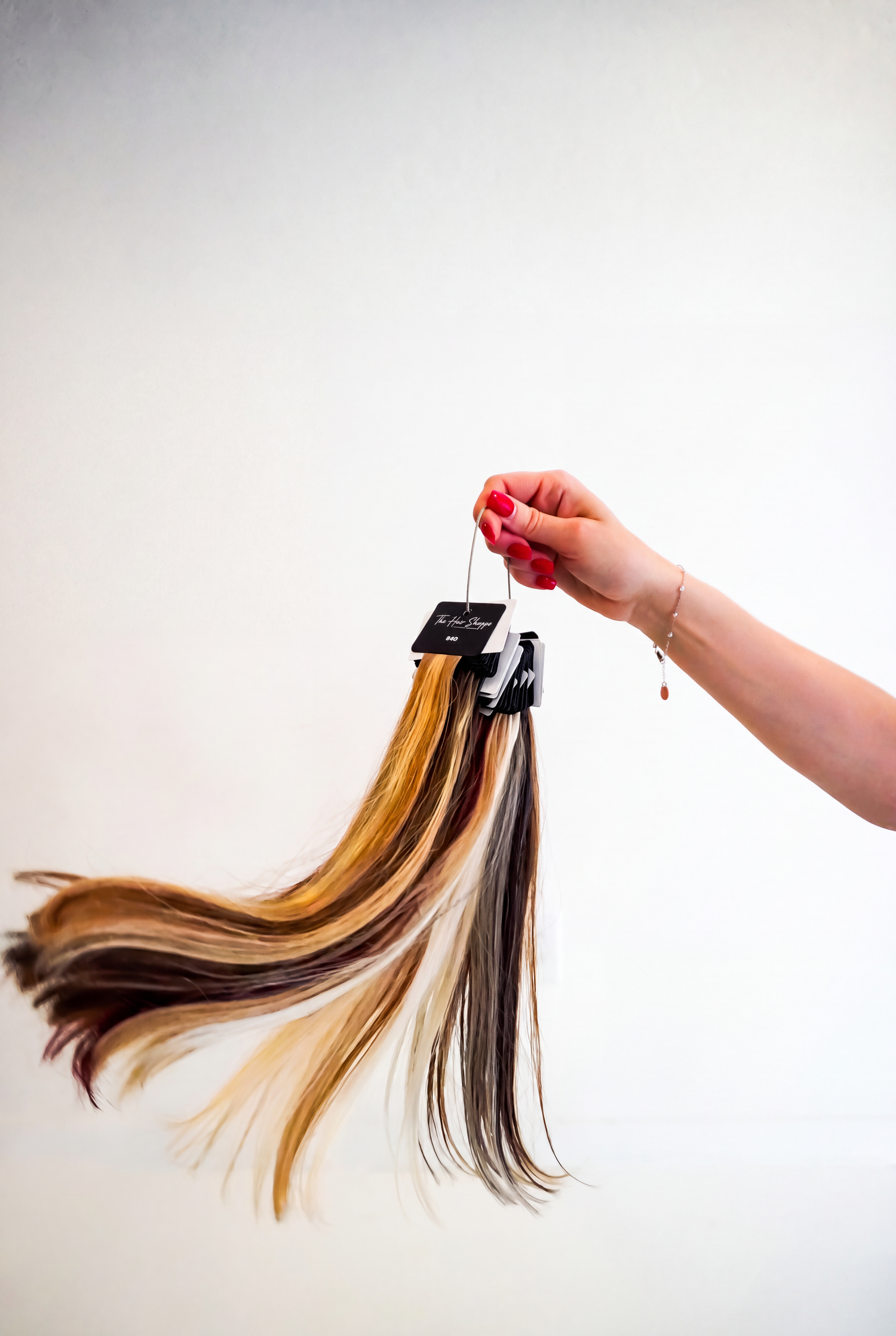 Hand with red nails holding a hair color swatch ring of blonde, brown, and black straight hair extensions against a white background.