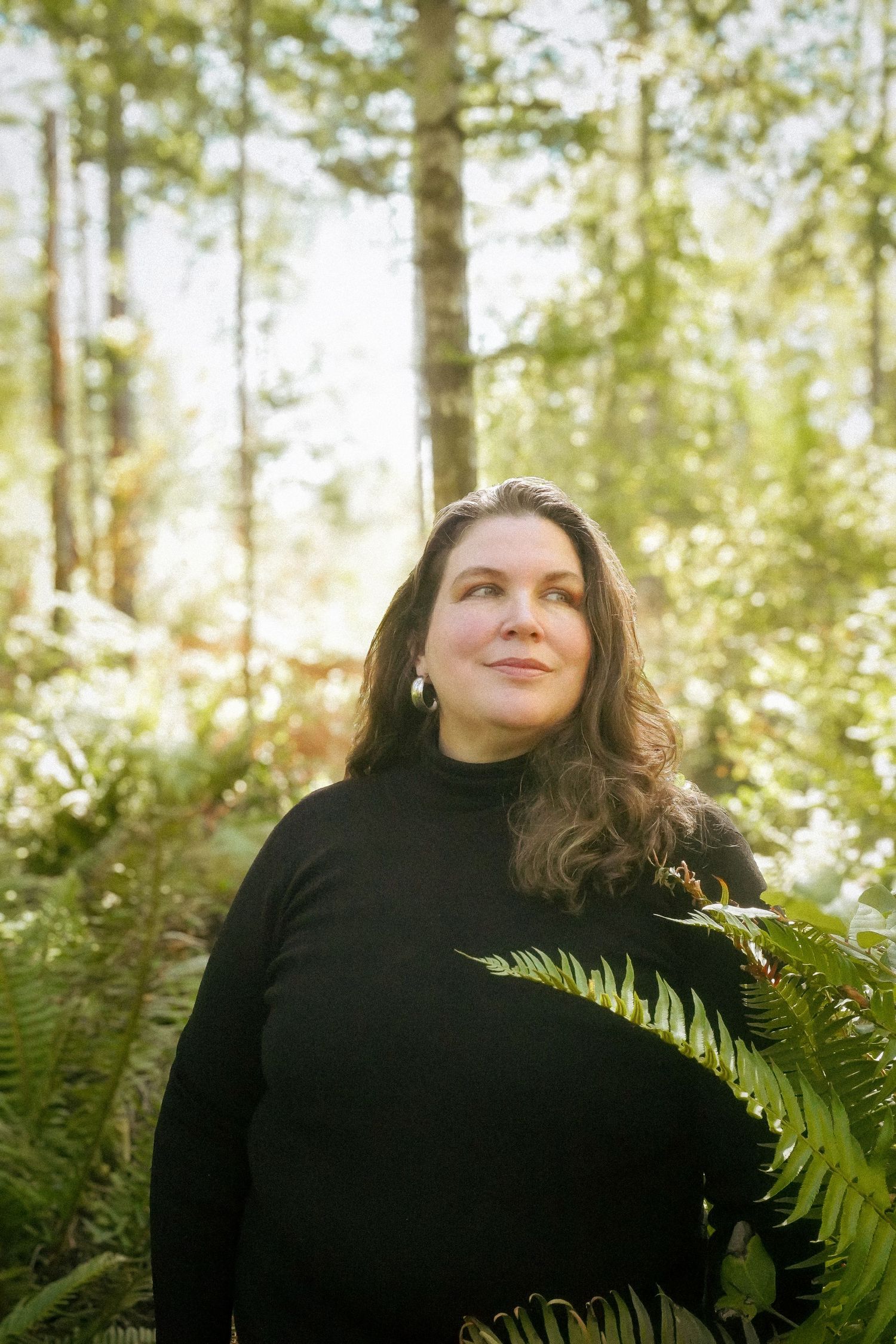 Woman in black turtleneck standing among ferns in a sunlit forest, gazing upward thoughtfully.