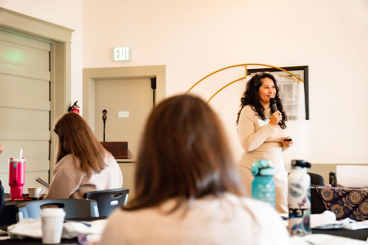 Woman with curly hair speaks into a microphone while presenting to seated attendees at a workshop in a bright meeting room.