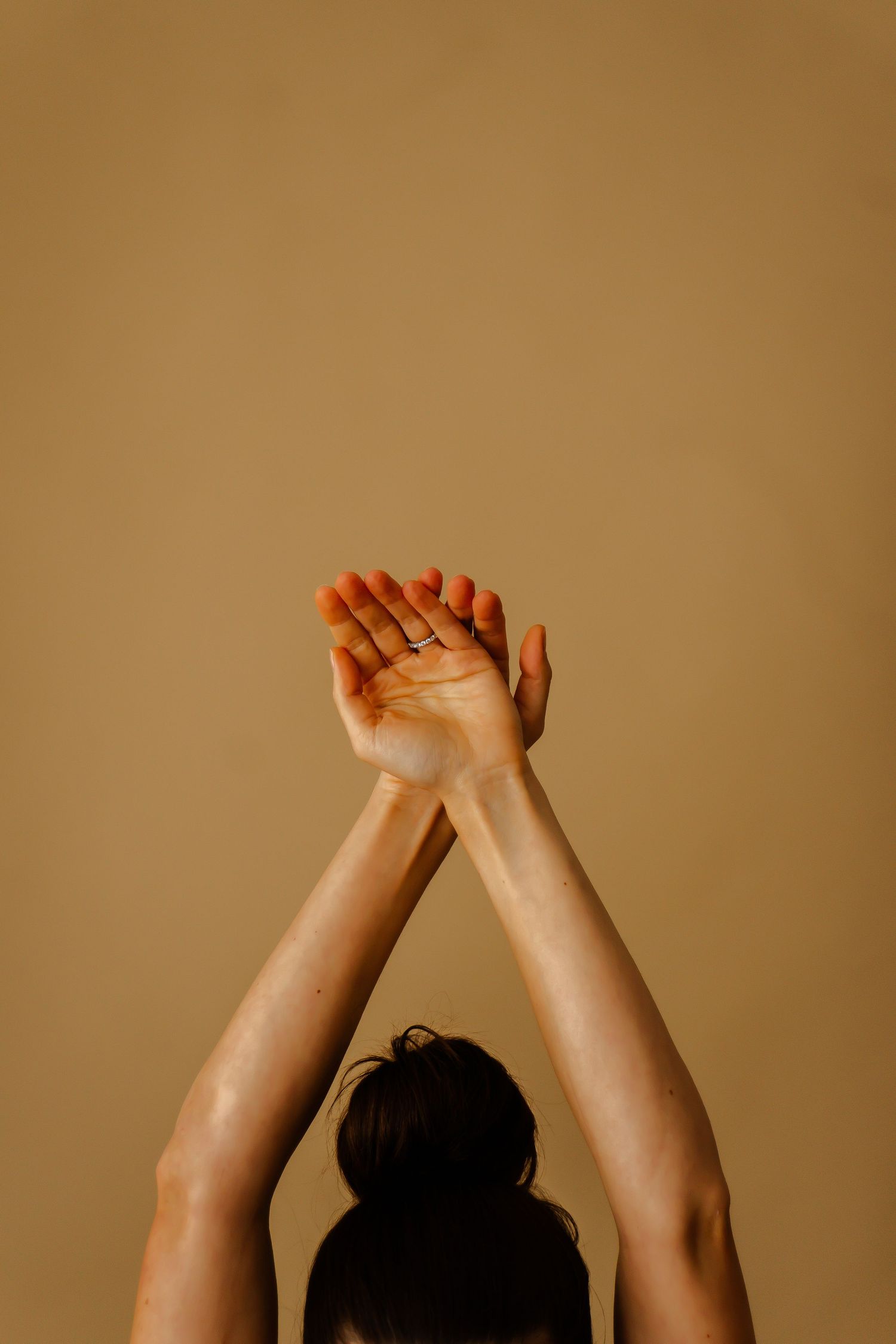 Woman with dark hair in a bun raises both arms overhead with palms open against a warm beige background.