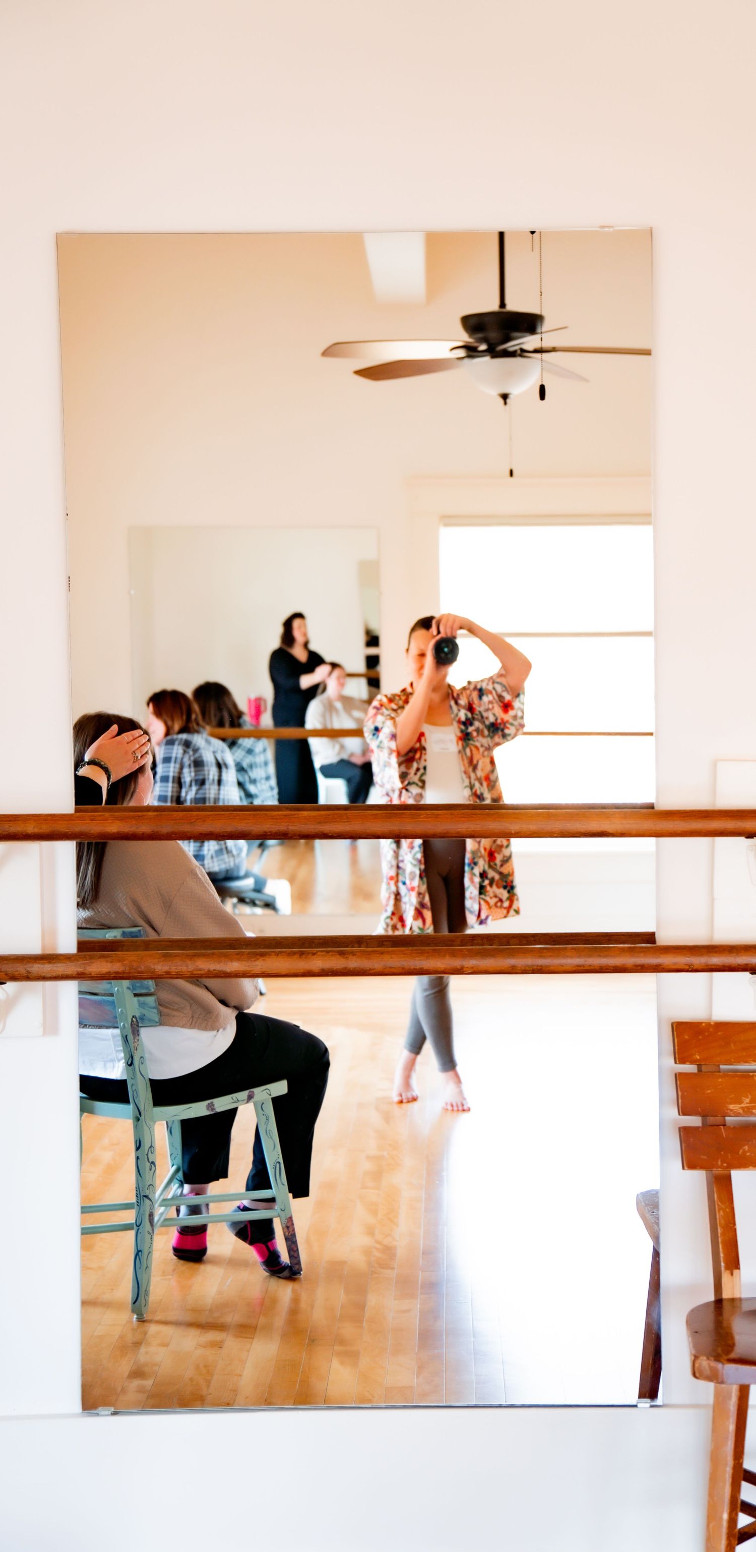 Mirror reflection in a bright dance studio with ballet barres, where a woman in a floral kimono takes a photo as others sit and chat nearby.